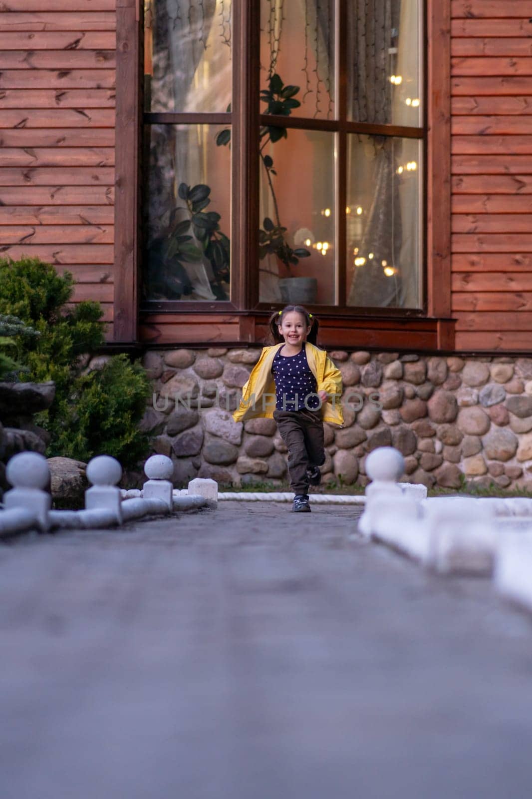Wonderful little lady with brown hair tied in ponytails, wearing bright yellow jacket running on street to camera with joyful smile shining on face, spending active walk near building. Vertical.