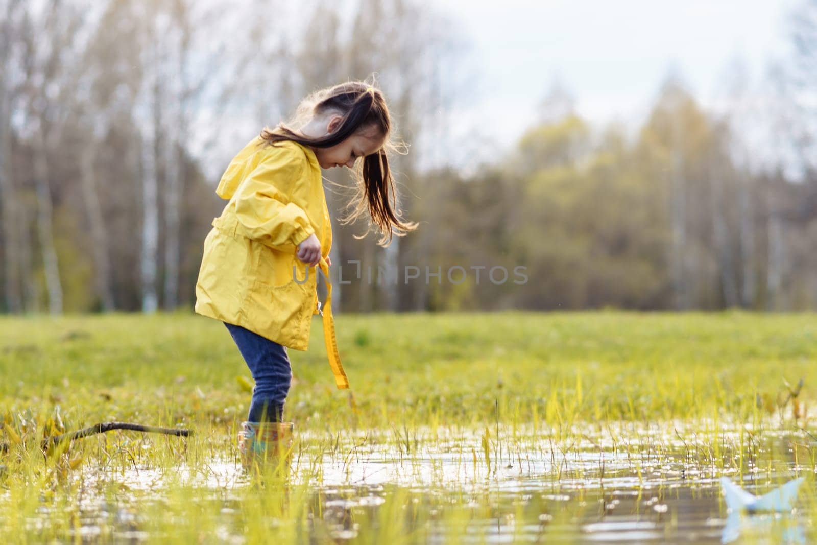 Curious little girl, wearing bright yellow jacket and rubber boots, standing in huge puddle on green site and looking down at water. Female child amusing to pool, enjoying leisure in nature.
