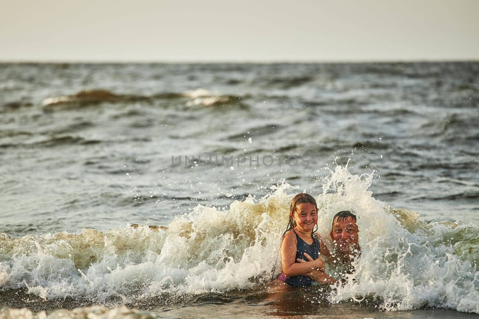 Little girl and her dad playing with waves in the sea. Family summer vacations. Kid playfully splashing with waves together with father. Jumping in sea waves. Summer vacation on the beach. Water splashes. Travel during summertime concept