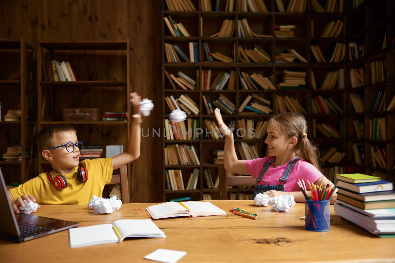 Cute funny children sitting at table and throwing crumpled paper sheets fooling around during online studying process. Naughty schoolkids enjoying fun time on virtual lesson
