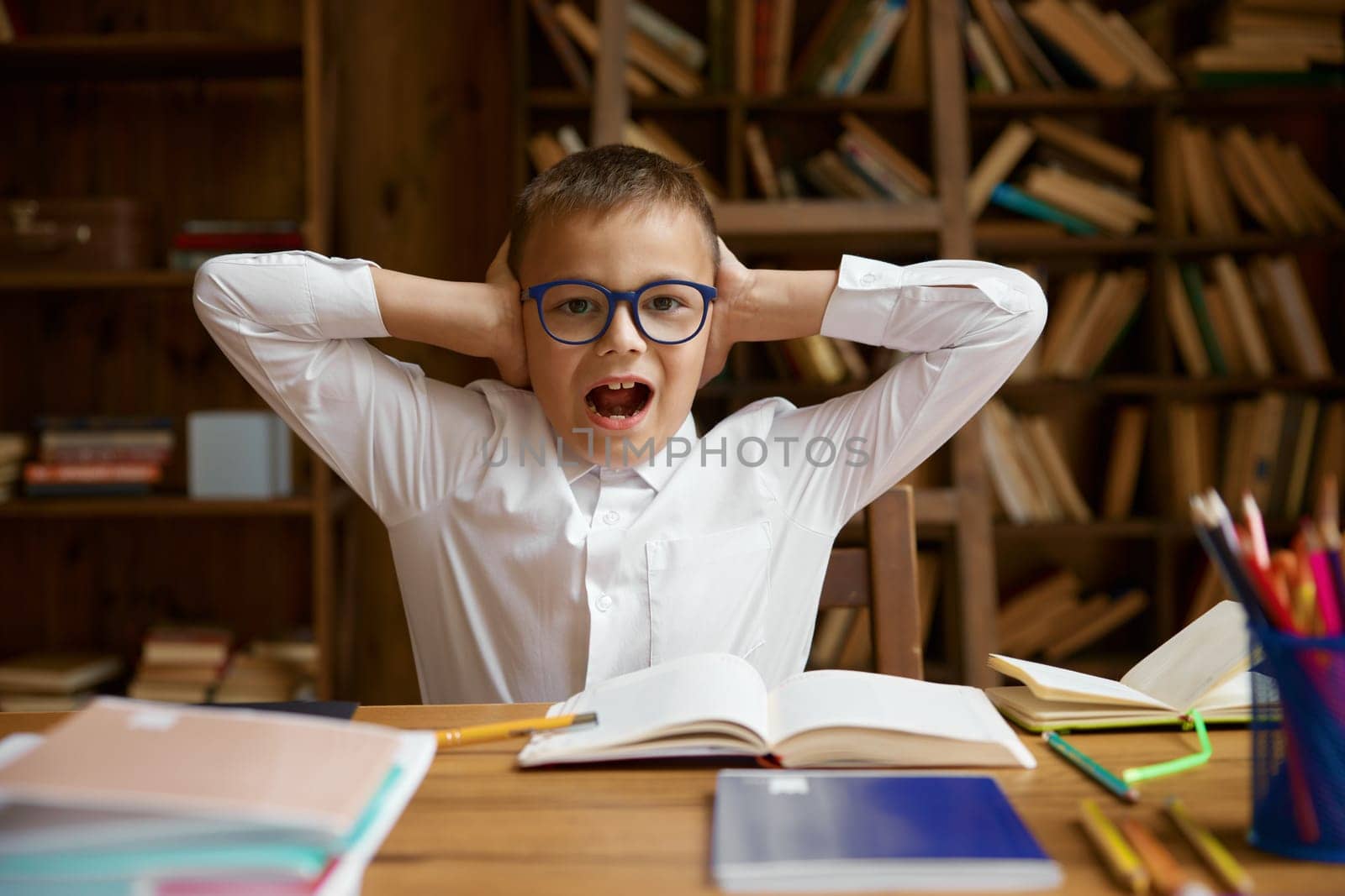 Upset mad schoolboy screaming covering ears while sitting at home table with homework. Portrait of cute little boy pupil yelling feeling annoyed suffering from noise