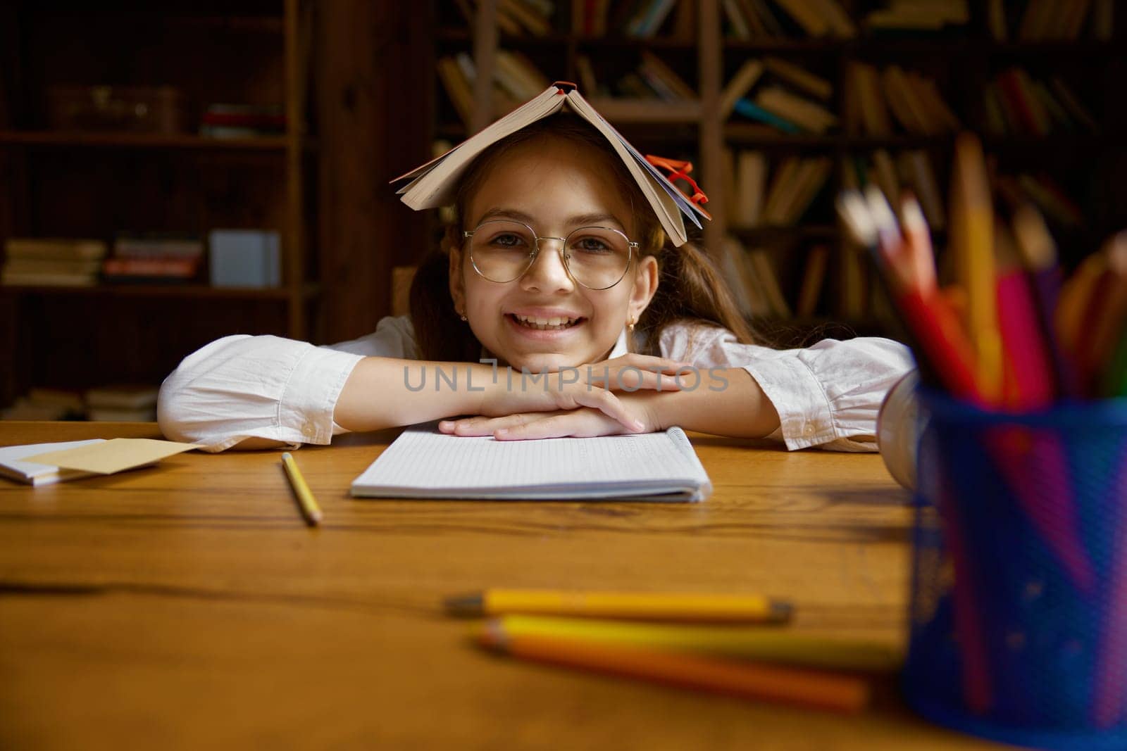Funny cute schoolgirl with book on head fooling around while sitting at table. Closeup portrait of elementary school pupil