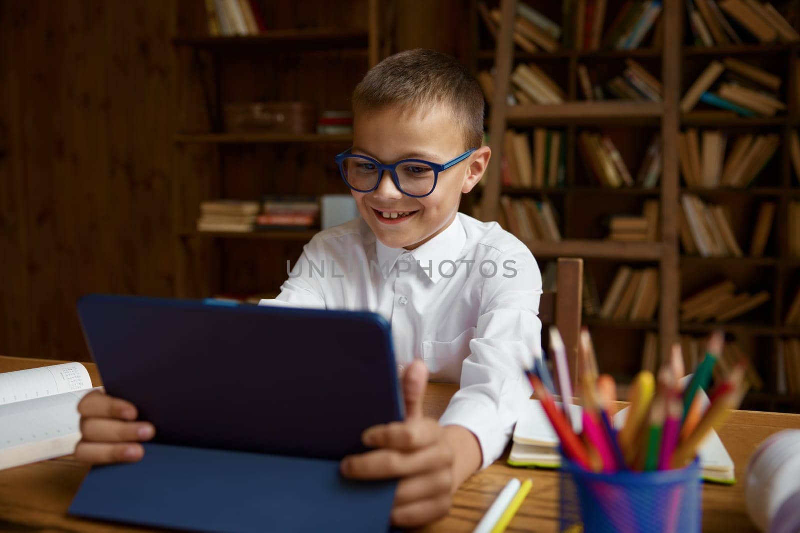 Cute smart boy schoolchild studying online using electronic device for watching funny video and learning while sitting at table, selective focus