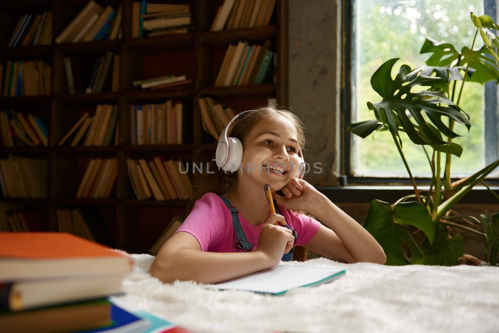 Cute little school girl wearing headphones enjoying school time having online lesson at home