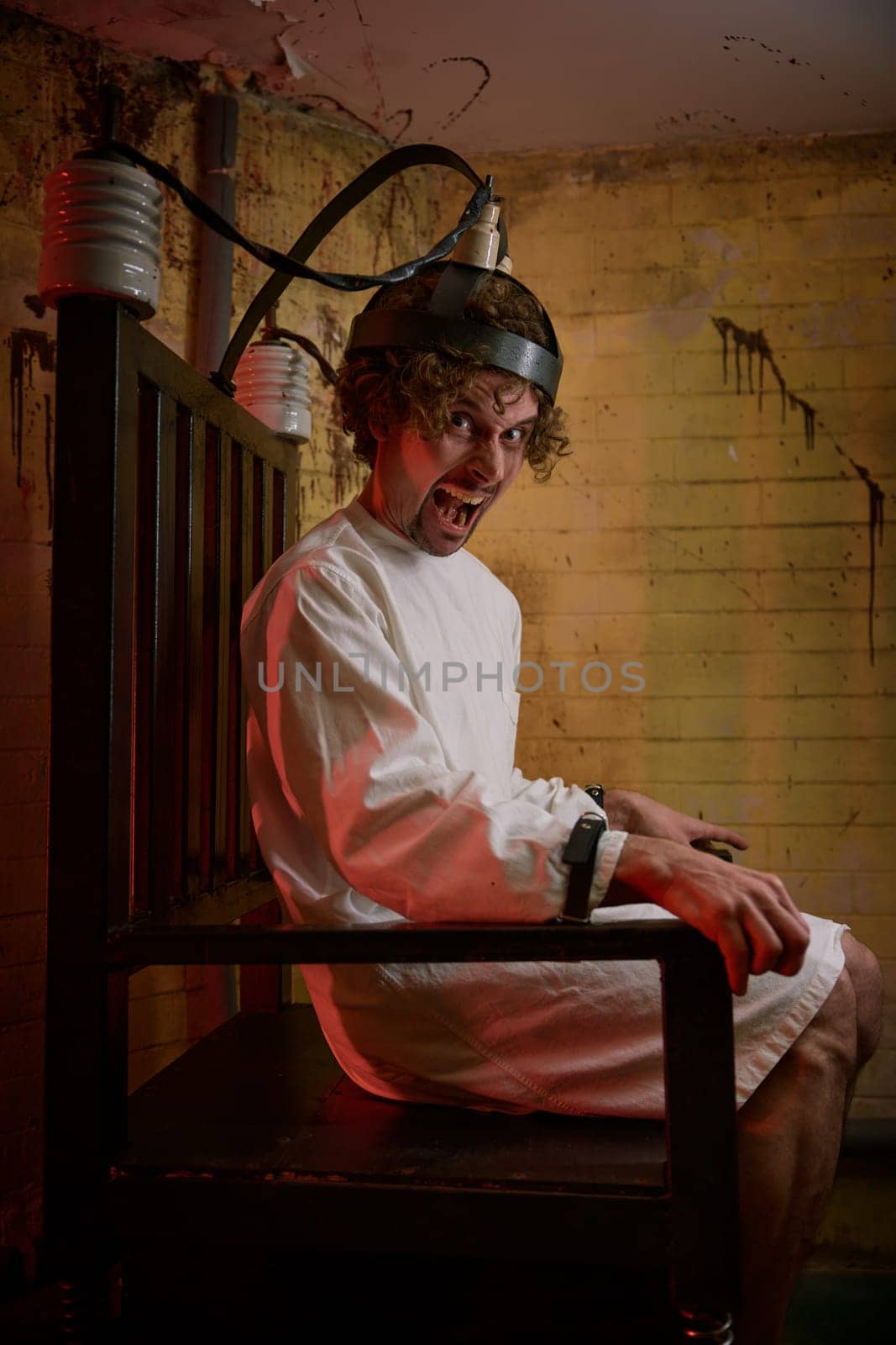 Crazy man screaming feeling madness and suffering from panic attack on electric chair at psychiatric hospital. Treatment mental ill patient with current