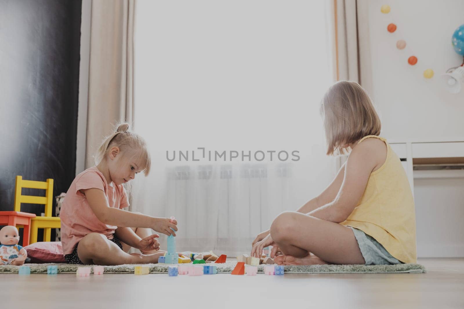 Children playing with colorful wooden toy building blocks, bricks. Kids sitting on floor at sunny white home bedroom or kindergarten, daycare playroom. Funny educational eco-friendly game for child