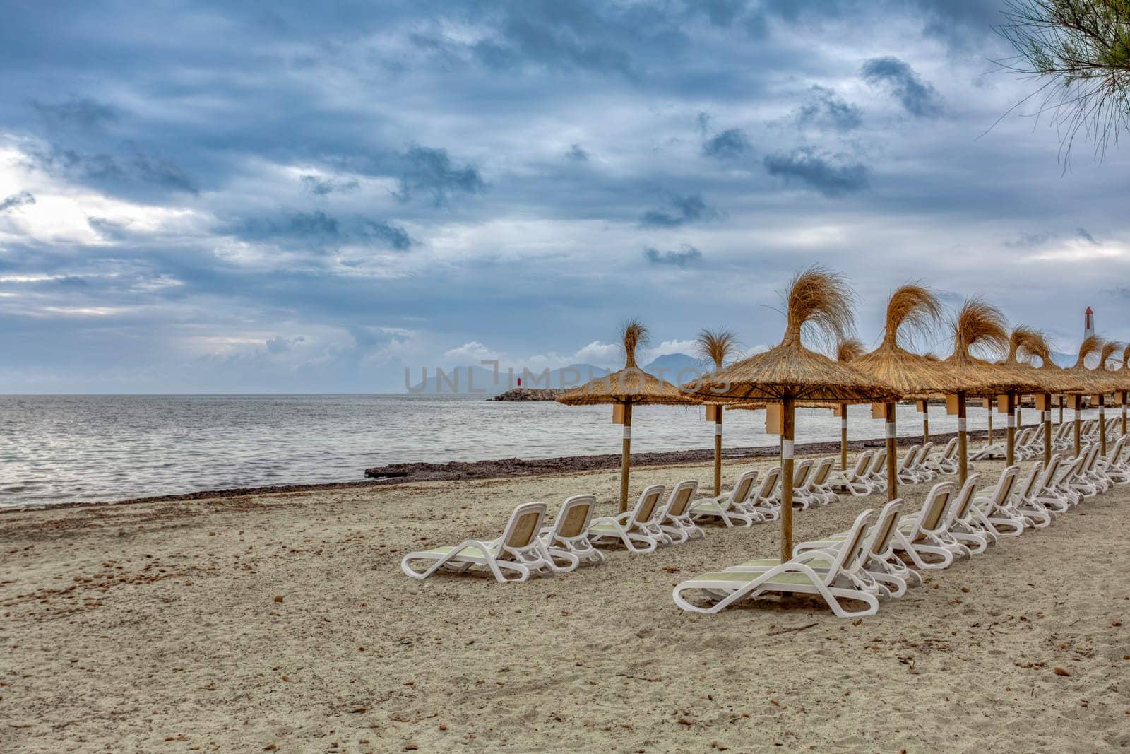 Can Picafort Beach with straw umbrellas and sun loungers, morning with no people, overcast cloudy weather. Can Picafort, Balearic Islands Mallorca Spain. Travel agency vacation concept.