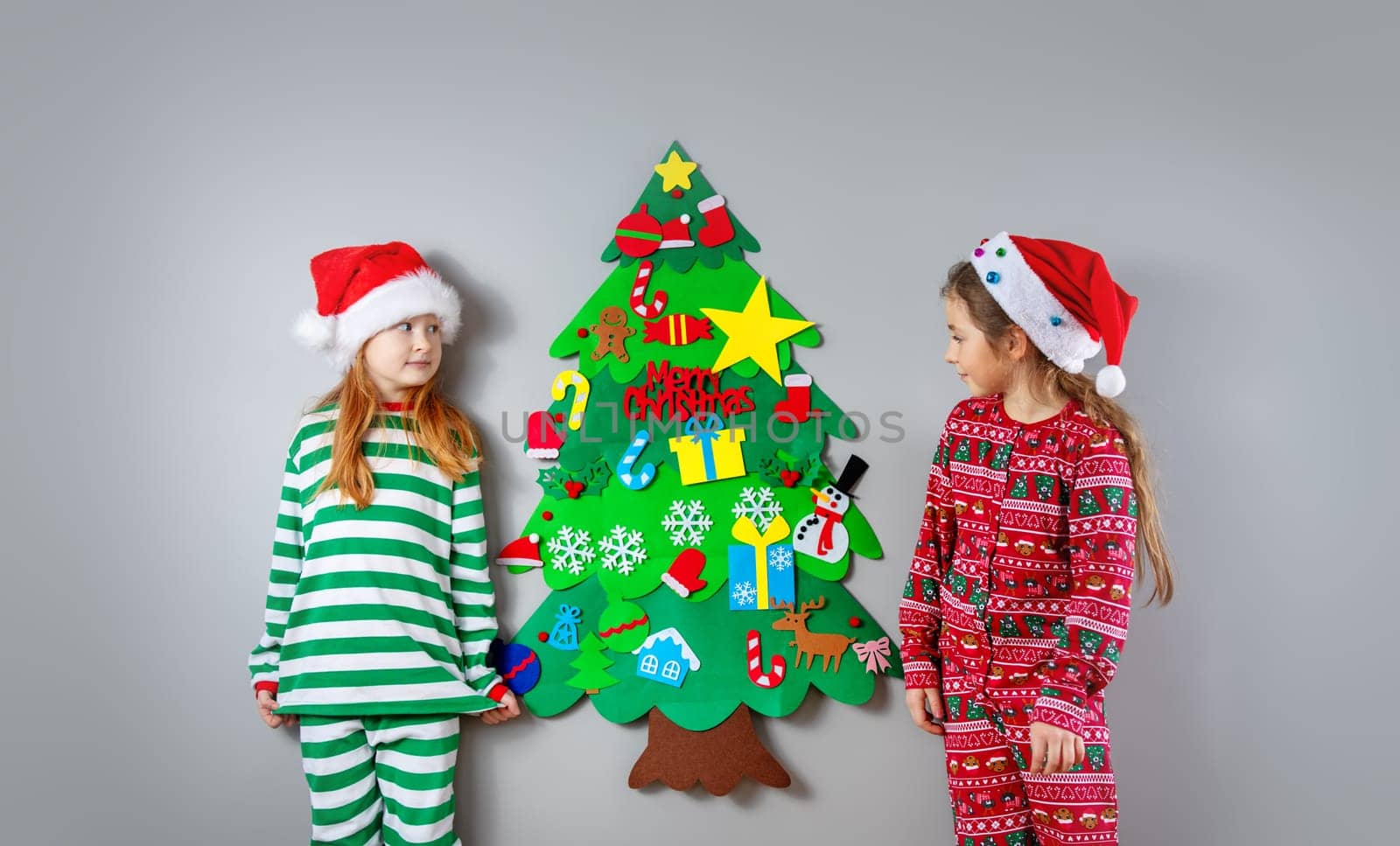 Two little girls decorating wall of the room by green toy fir Christmas tree. Concept of the Xmas and New Year.