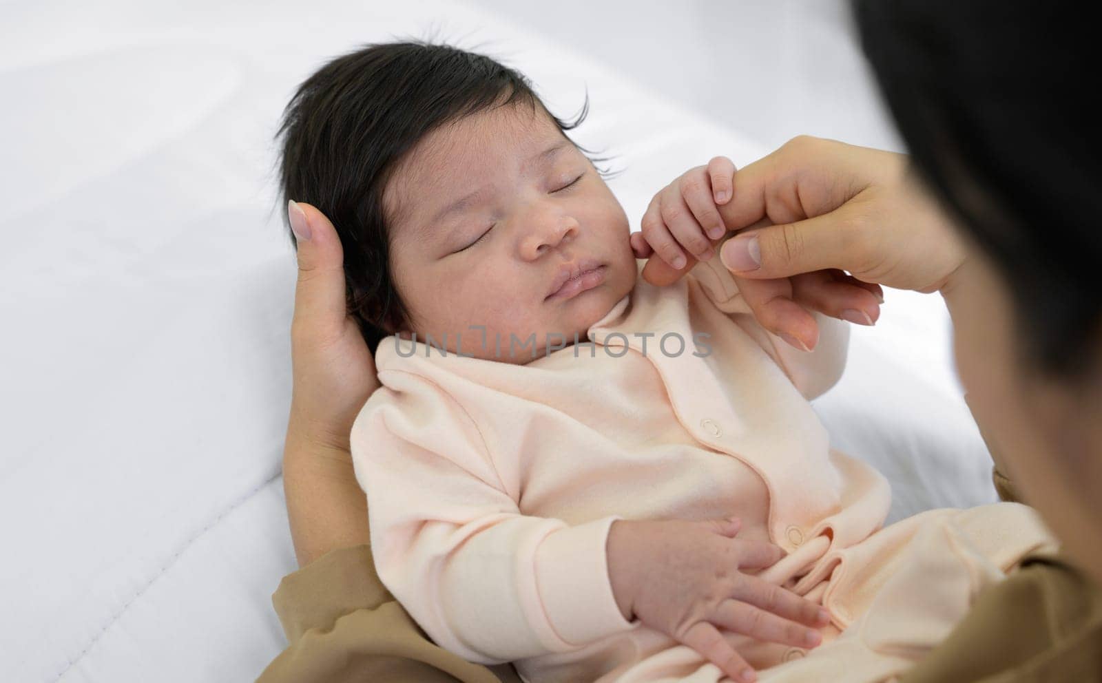Cute newborn girl or boy 1 month sleeping on mom hand at home. Mother sitting on white bed and gently touching cheek of infant baby daughter or son with love. Care, love and mothers day concept.
