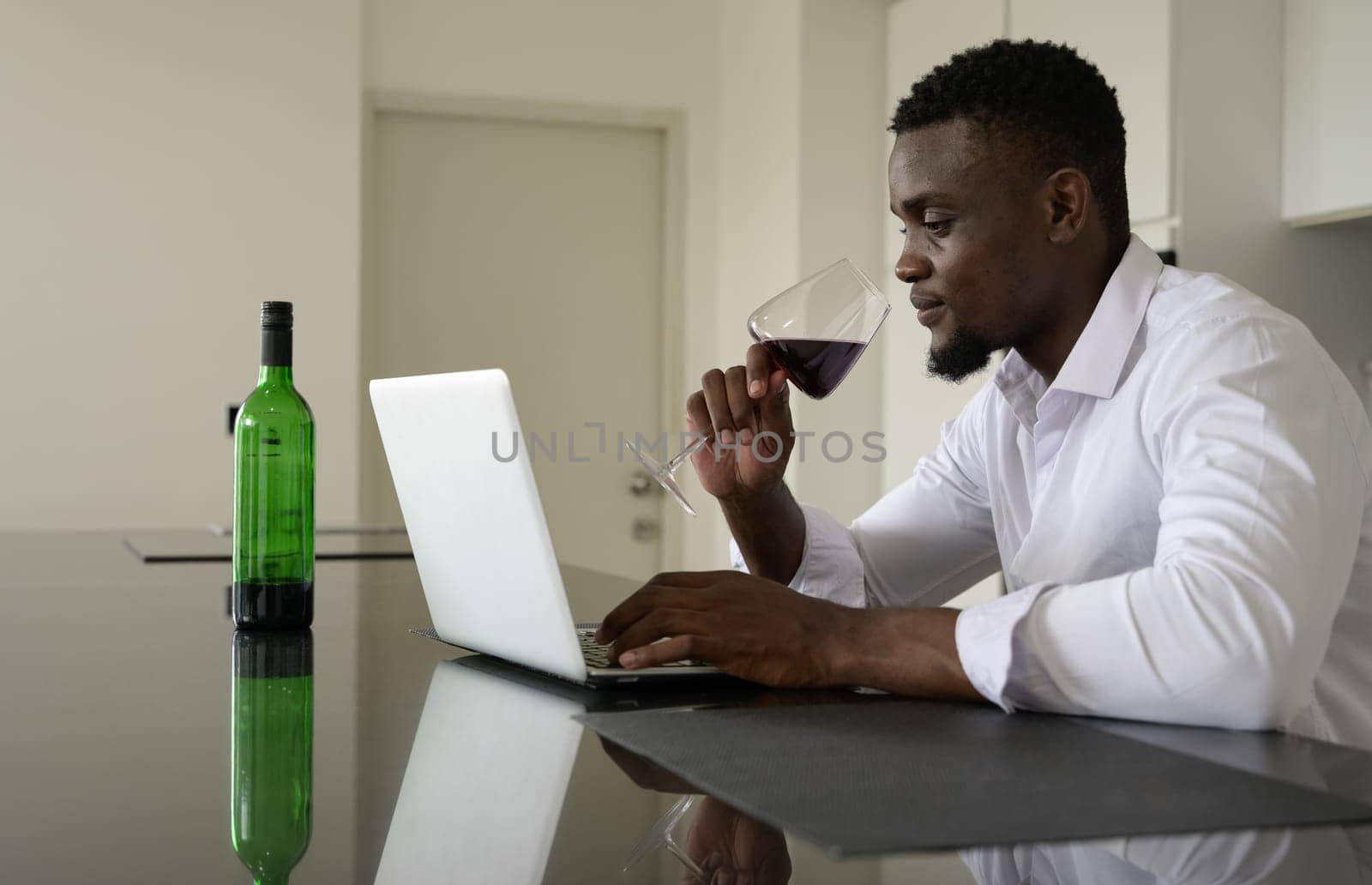 Businessman in white shirt drinking red wine and using laptop in kitchen. Working from home concept.