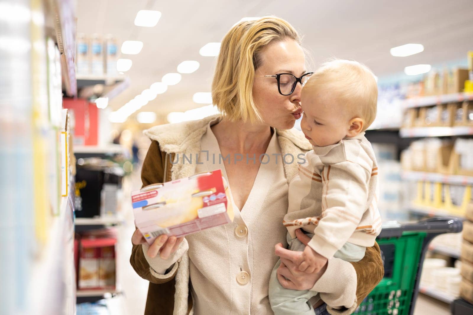 Caucasian mother shopping with her infant baby boy child choosing products in department of supermarket grocery store