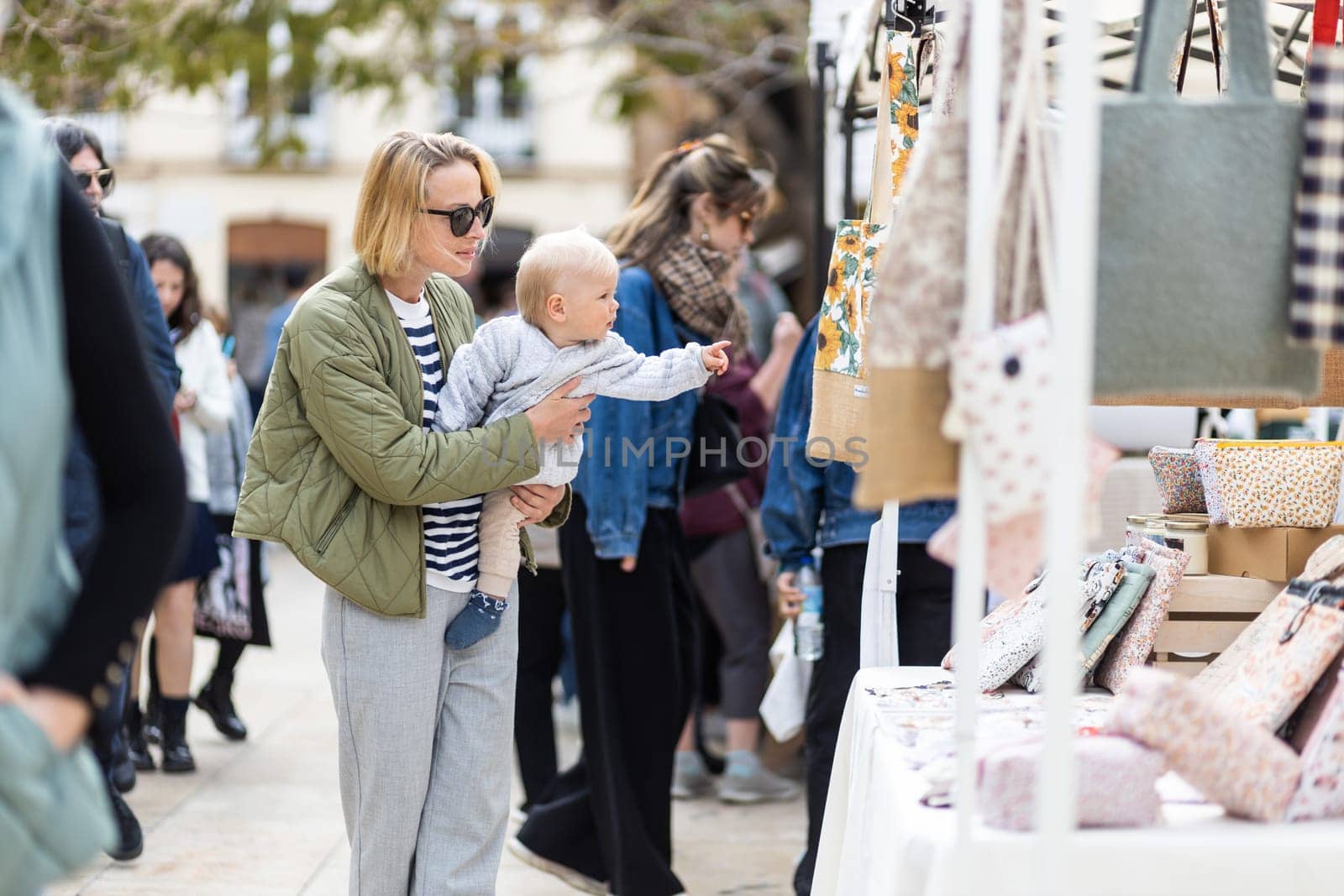 Mother walking carrying his infant baby boy child in crowd of people wisiting sunday flea market in Malaga, Spain.