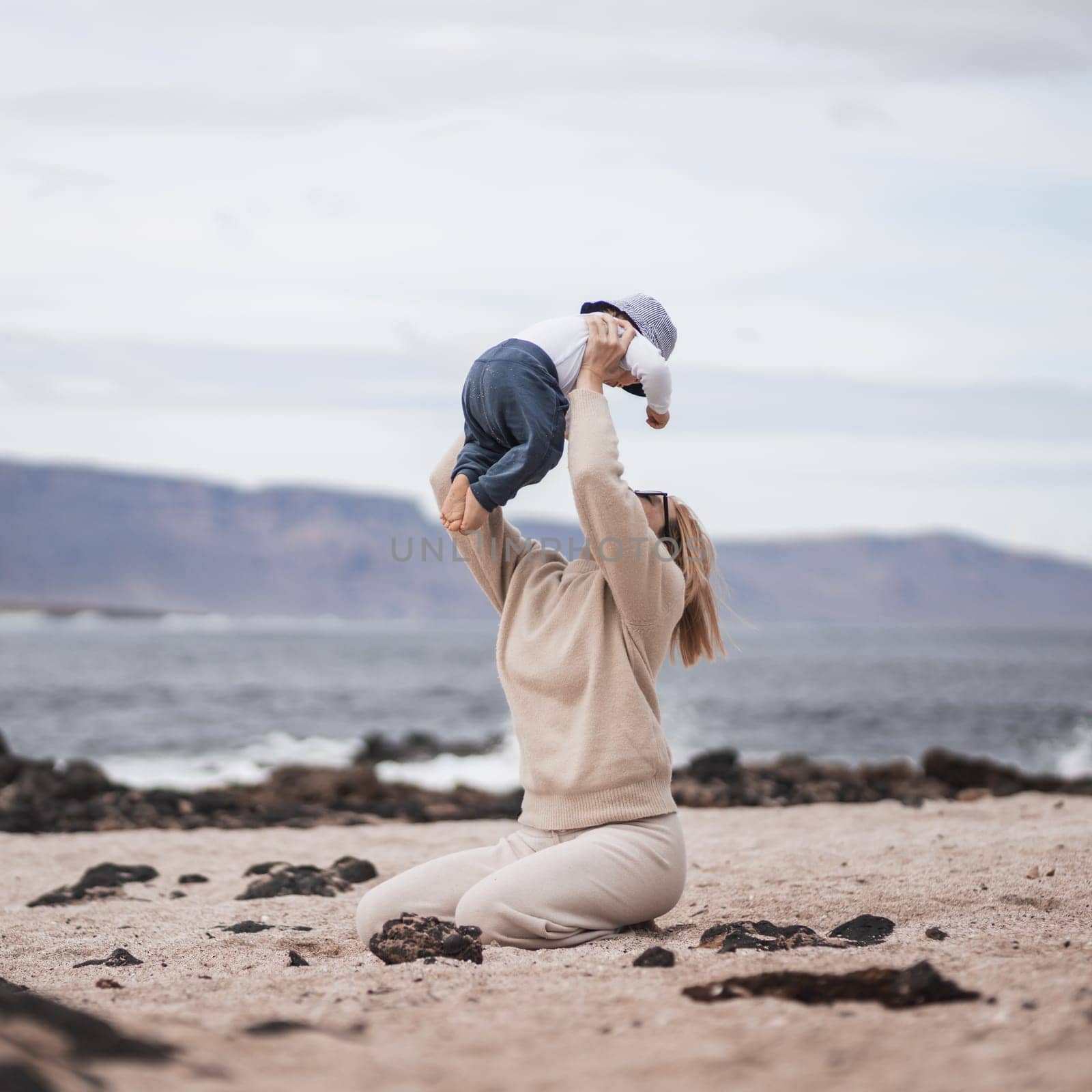 Mother enjoying winter vacations holding, playing and lifting his infant baby boy son high in the air on sandy beach on Lanzarote island, Spain. Family travel and vacations concept.
