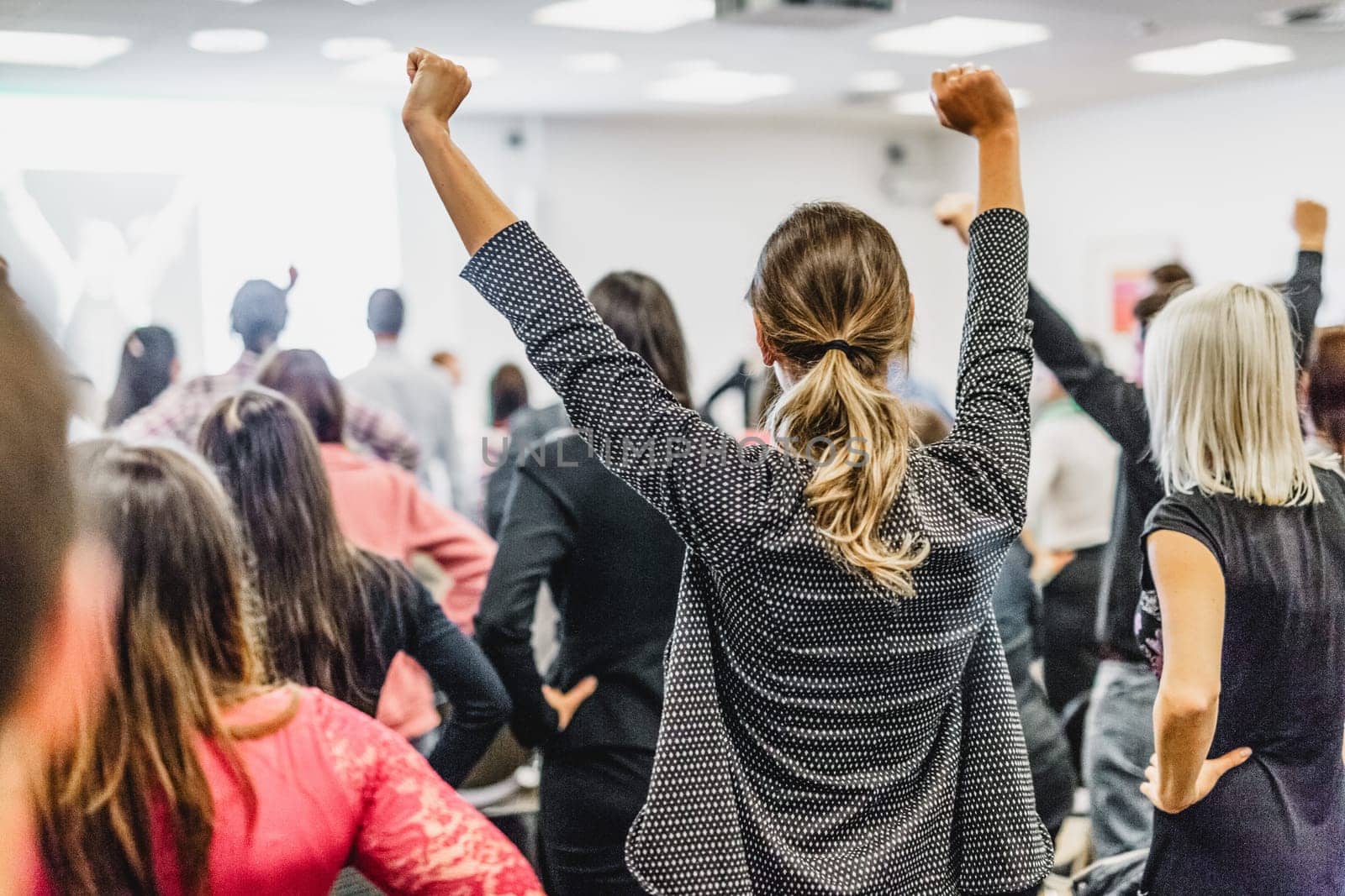 Life coaching symposium. Speaker giving interactive motivational speech at business workshop. Rear view of unrecognizable participants feeling empowered and motivated, hands raised high in air.