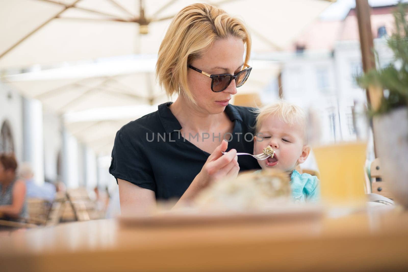 Young caucasian blonde mother spoon feeding her little infant baby boy child outdoors on restaurant or cafe terrace in summer.