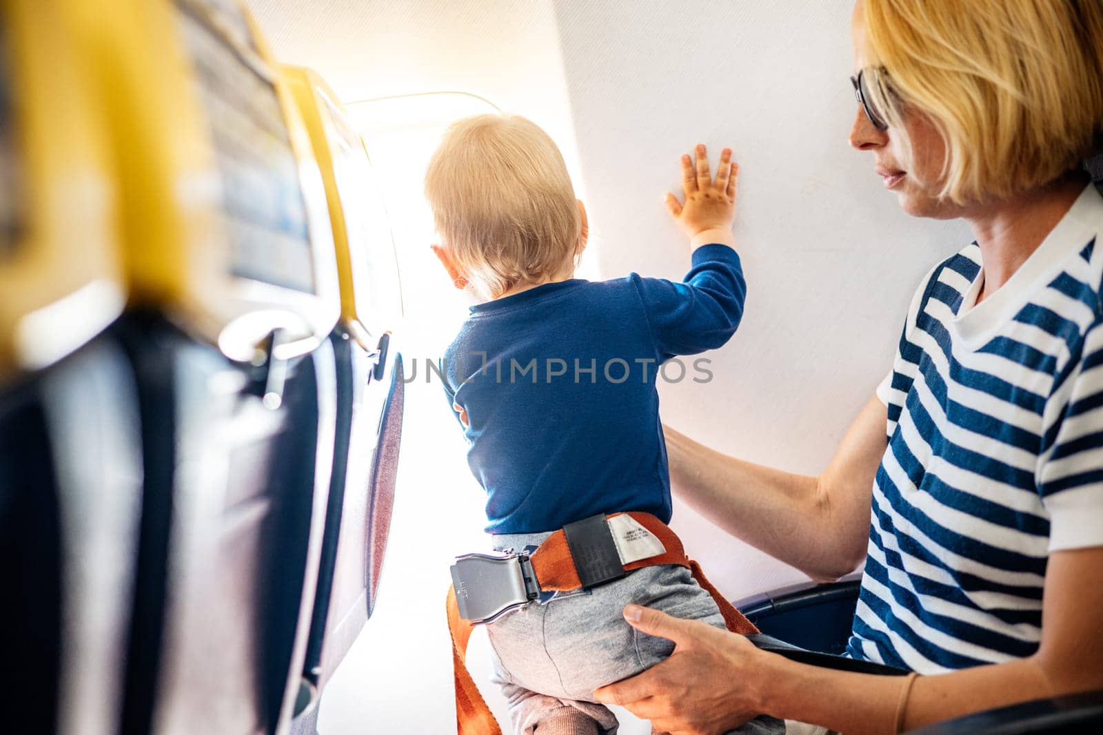 Mom and child flying by plane. Mother holding and playing with her infant baby boy child in her lap during economy comercial flight. Concept photo of air travel with baby. Real people