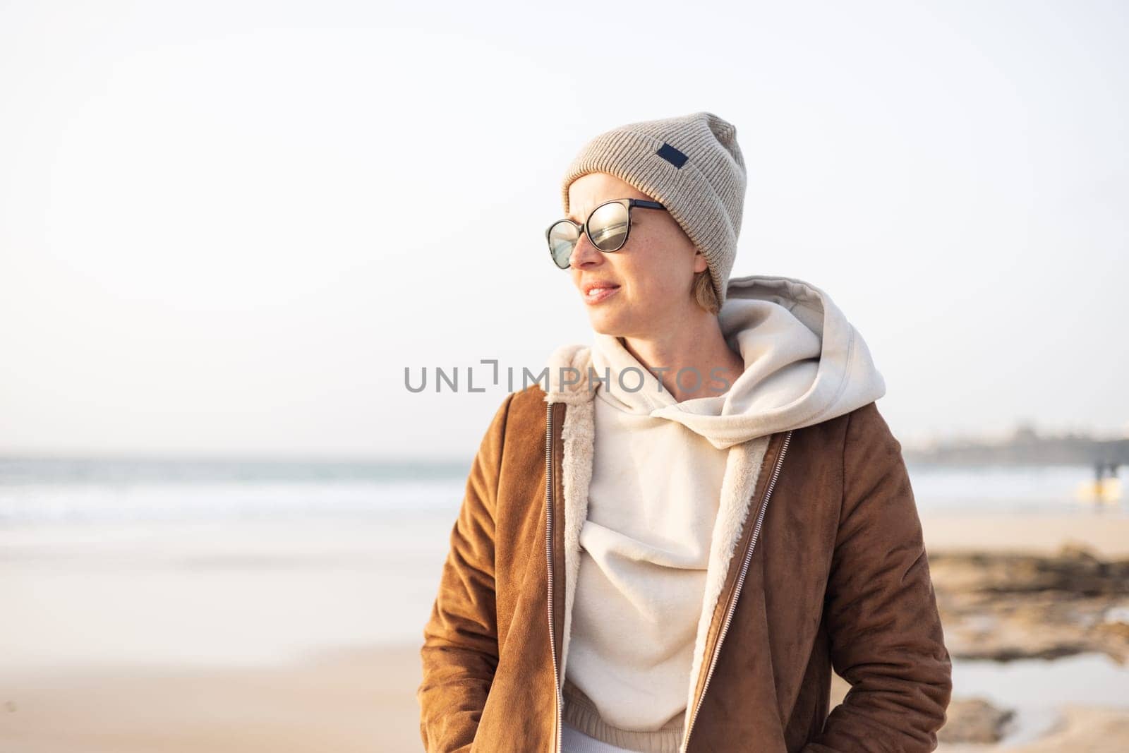 Portrait of young stylish woman wearing brown padded jacket, hoodie, wool cap and sunglasses on long sandy beach in spring