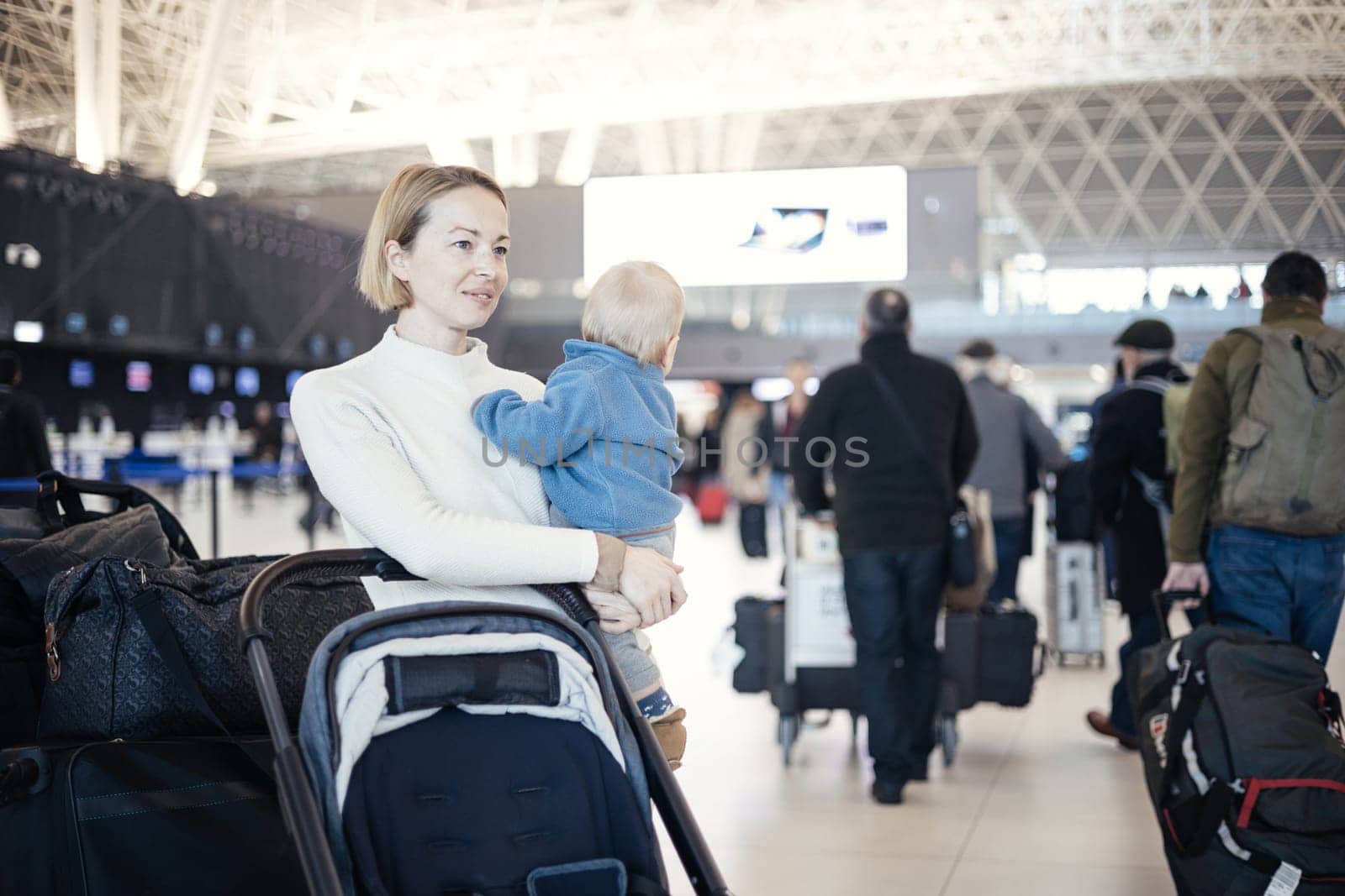 Motherat travelling with his infant baby boy child, walking, pushing baby stroller and luggage cart at airport terminal station. Travel with child concept