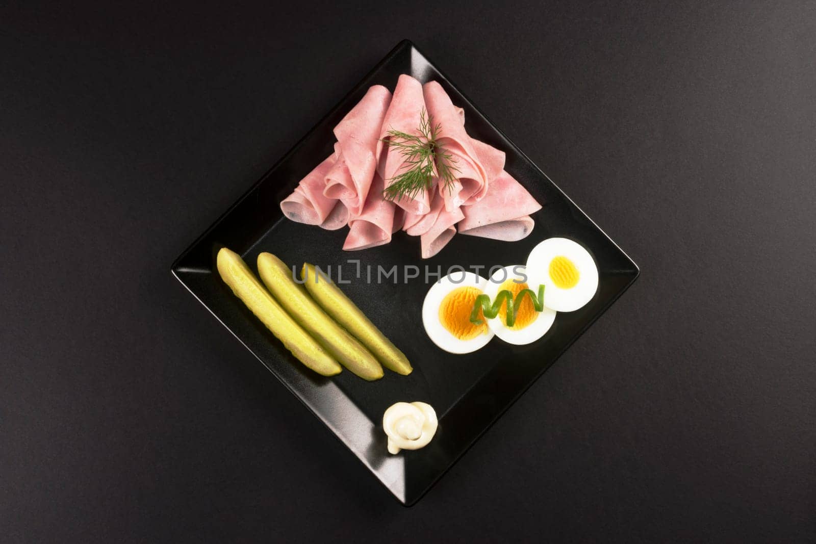 Elegant cold plate appetizer with ham, eggs and pickle on black plate on dark background.