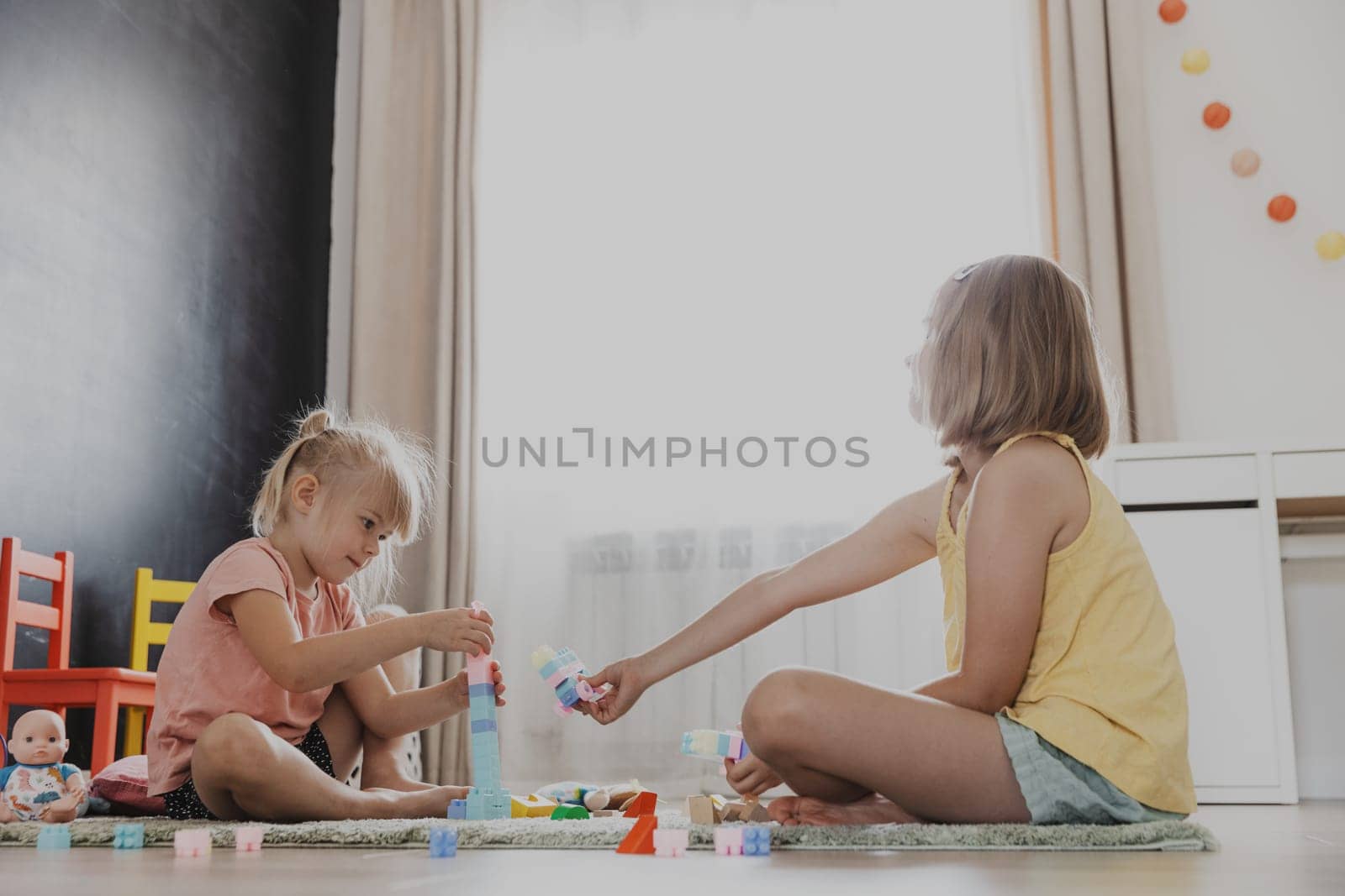 Children playing with colorful constructor building blocks, bricks. Kids sitting on floor indoors at sunny white home bedroom or kindergarten, play center. Funny educational child eco-friendly game