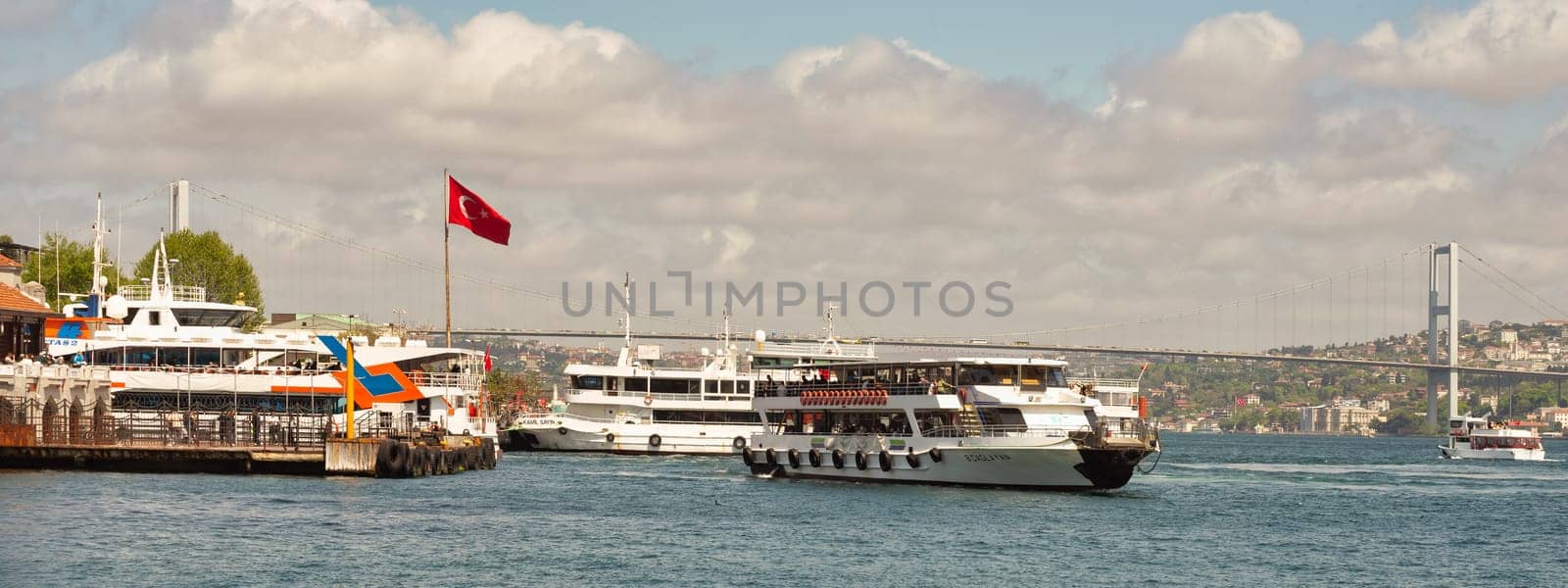 Istanbul, Turkey - May 7, 2023: Boats float peacefully on the Bosphorus Strait near Besiktas ferry terminal. The iconic Bosphorus Bridge towers in the background. The sky is cloudy in a spring day