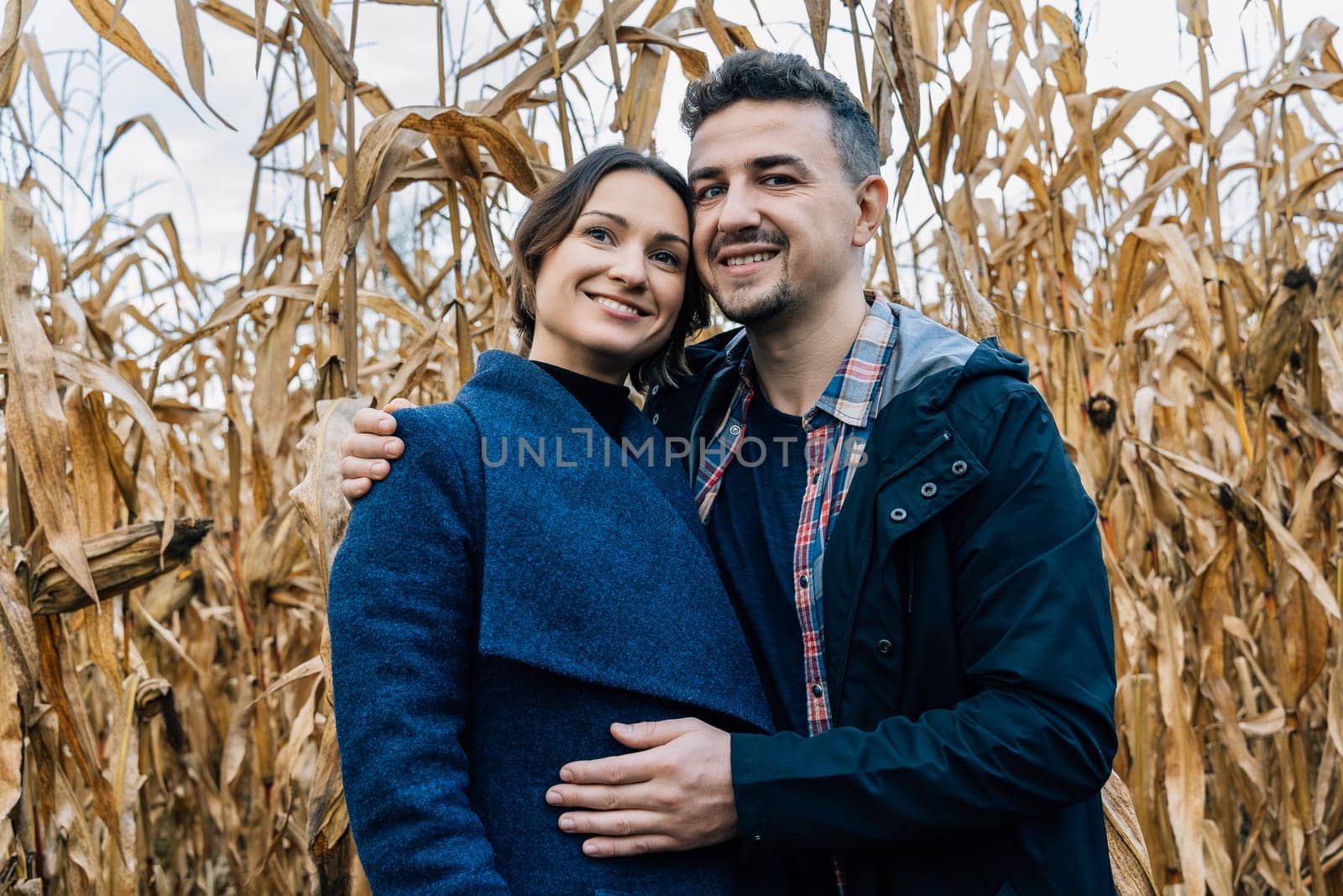 A happy couple in love stands in autumn near a cornfield. A man in a shirt and jacket hugs a woman in a coat.
