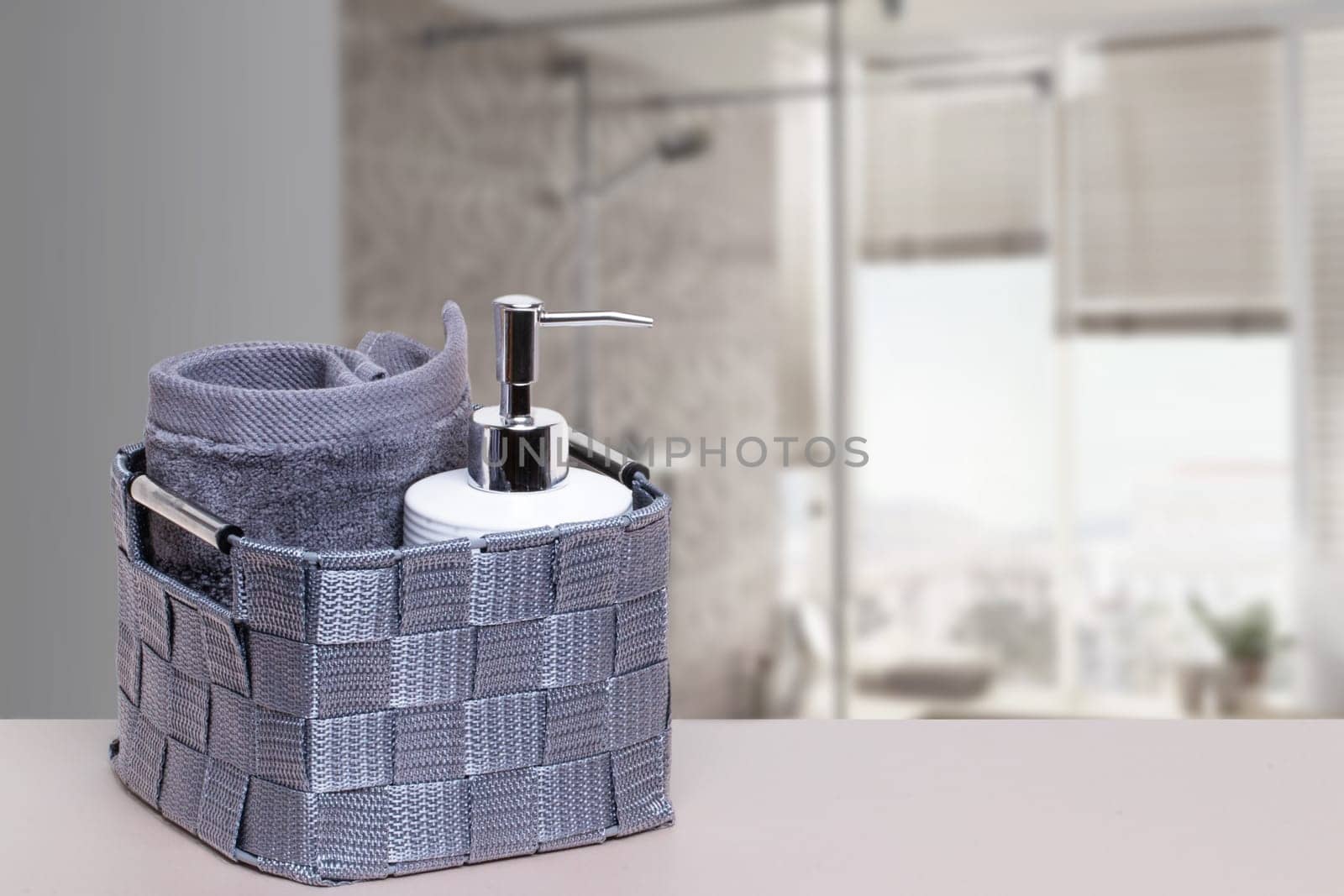 Basket with a fresh soft terry bath towel and a soap bottle on a white table over blurred bath background with copy space. For your product display montage.