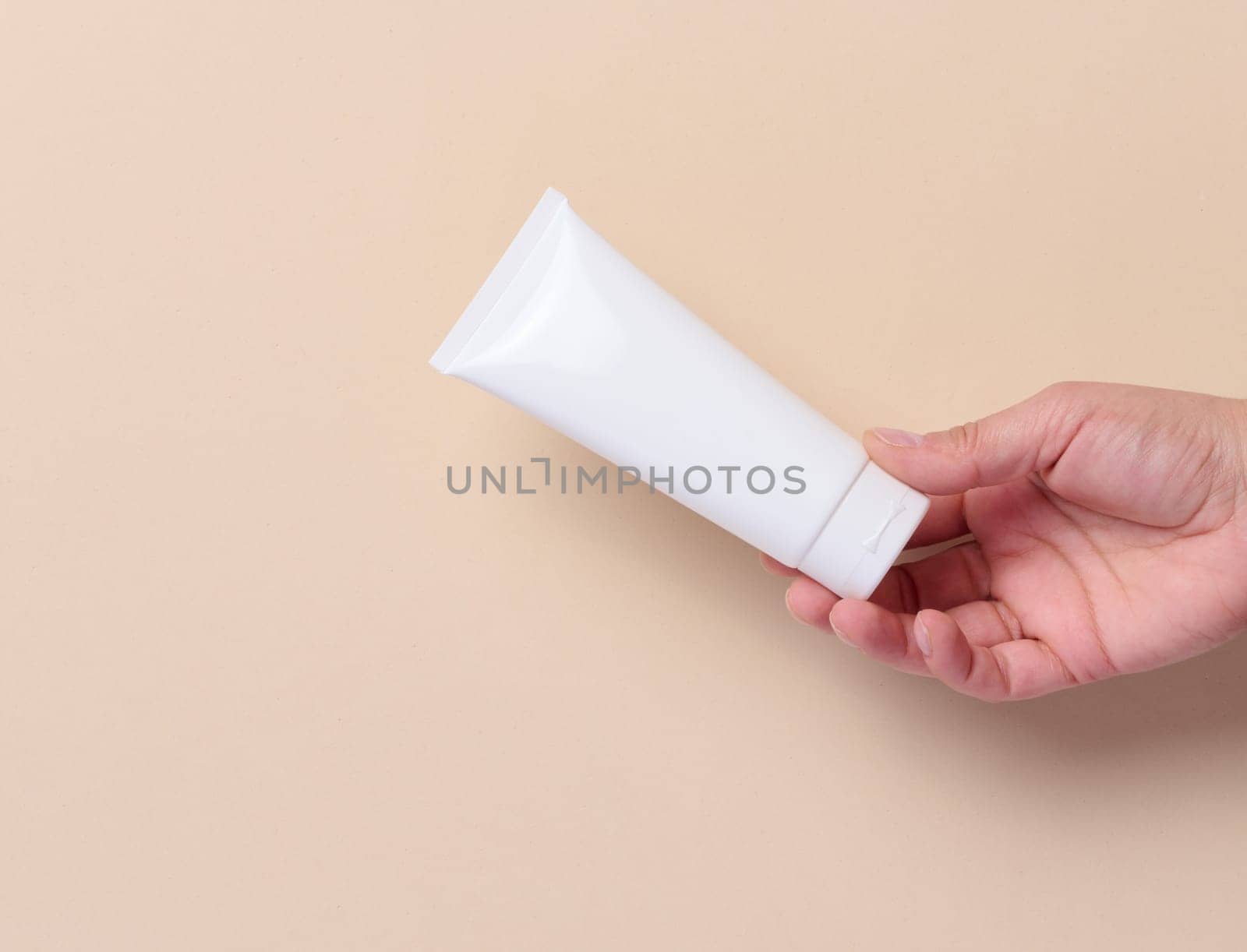A woman's hand holds a white plastic tube for cream, toothpaste on a beige background
