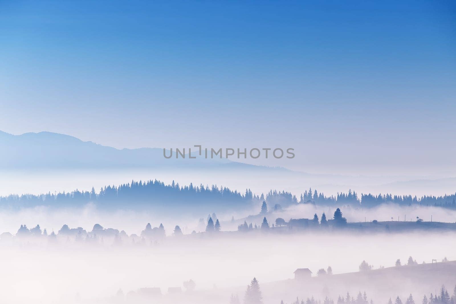 Clear blue sky and clouds of fog moving over alpine village on hills. Tranquil sunrise landscape. Sea of mist in valley. Autumn sunny morning in mountains. Carpathian range, Ukraine