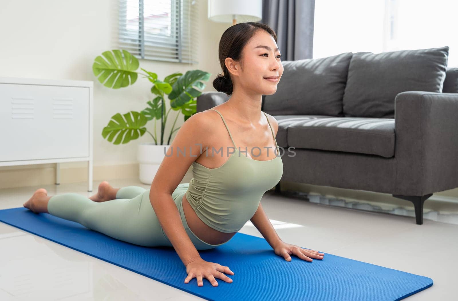 Young female practicing morning yoga routine at home, lying in Cobra pose or Bhujangasana to stretches back bend and spine on blue mat in living room. Healthy life and fitness training workout at home concept