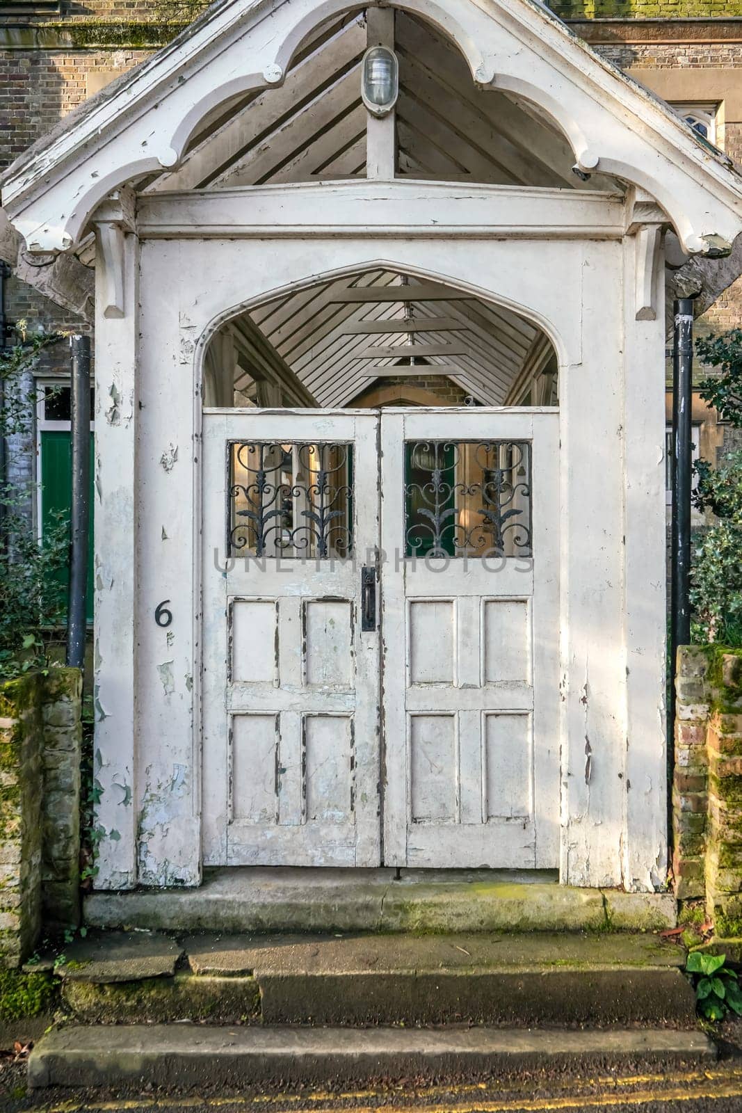 Sun shines on old white double door with number 6, leading to house terrace, bottom part dirty, paint peeling visible in places.