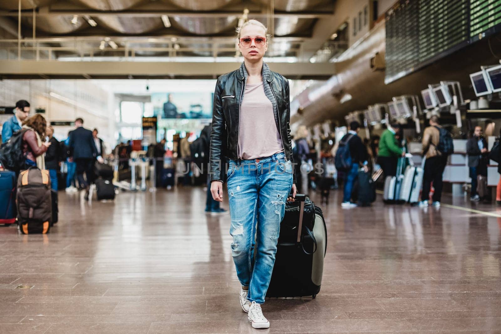 Casually dressed young stylish female traveler walking the airport terminal hall dragging suitcase and a handbag behind her. Blurred background. Can also be used as railway, metro, bus station.