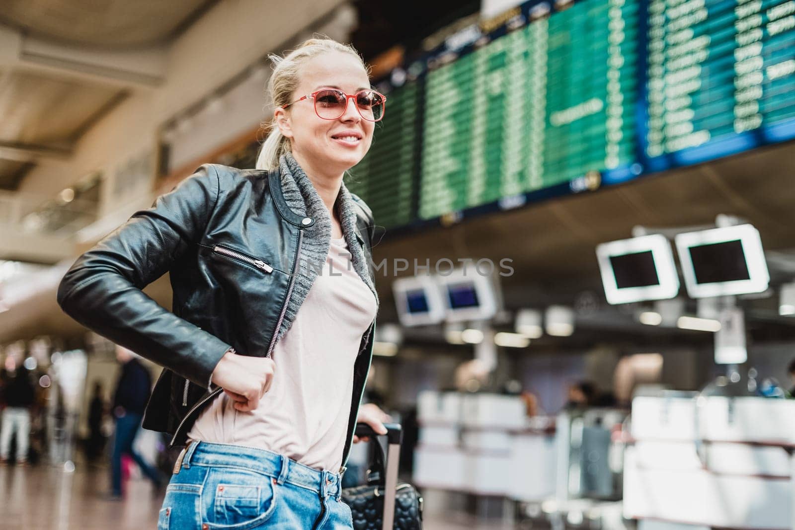 Casually dressed young stylish female traveller checking a departures board at the airport terminal hall in front of check in couters. Flight schedule display blured in the background. Focus on woman.