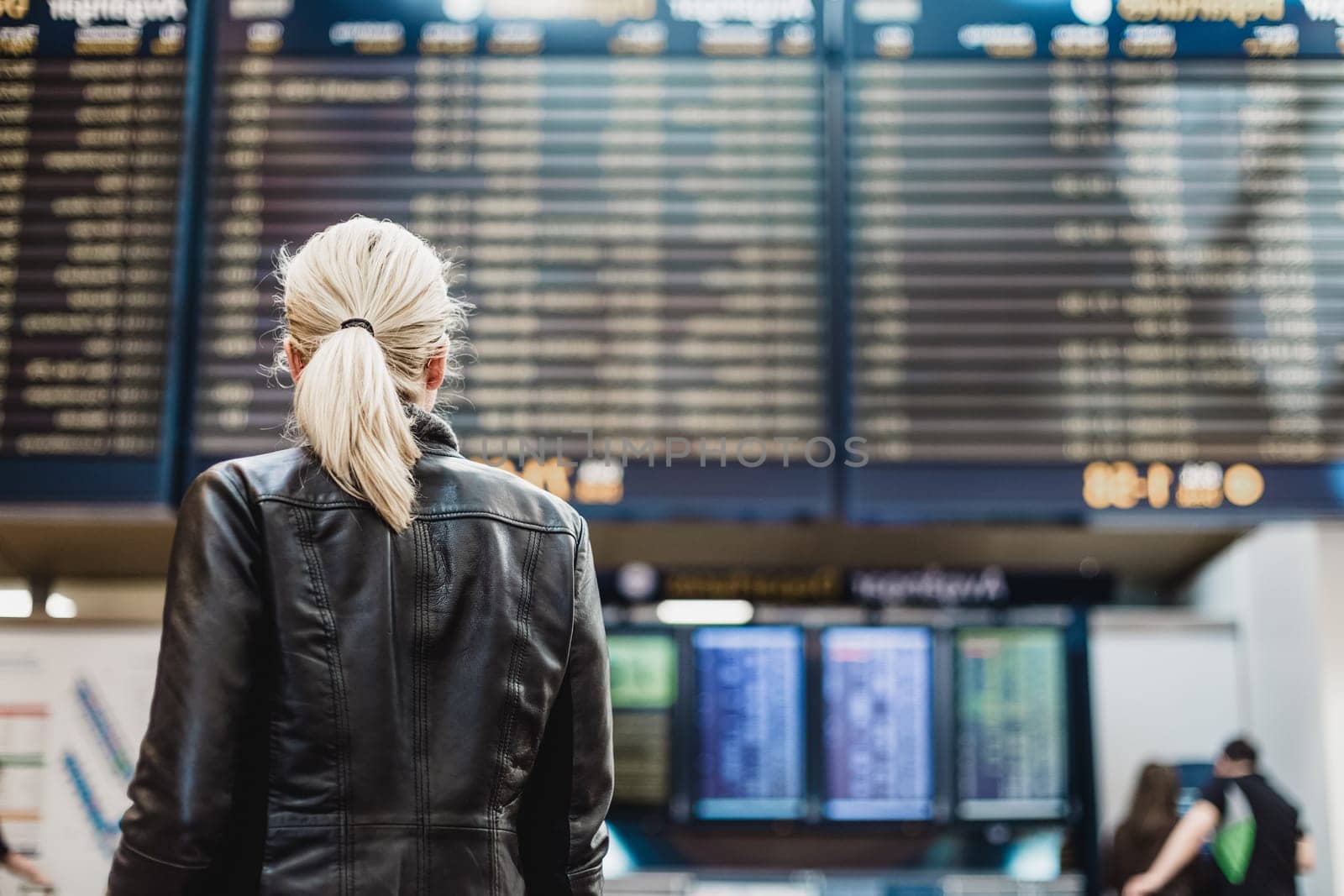 Casually dressed young stylish female traveller checking a departures board at the airport terminal hall in front of check in couters. Flight schedule display blured in the background. Focus on woman.