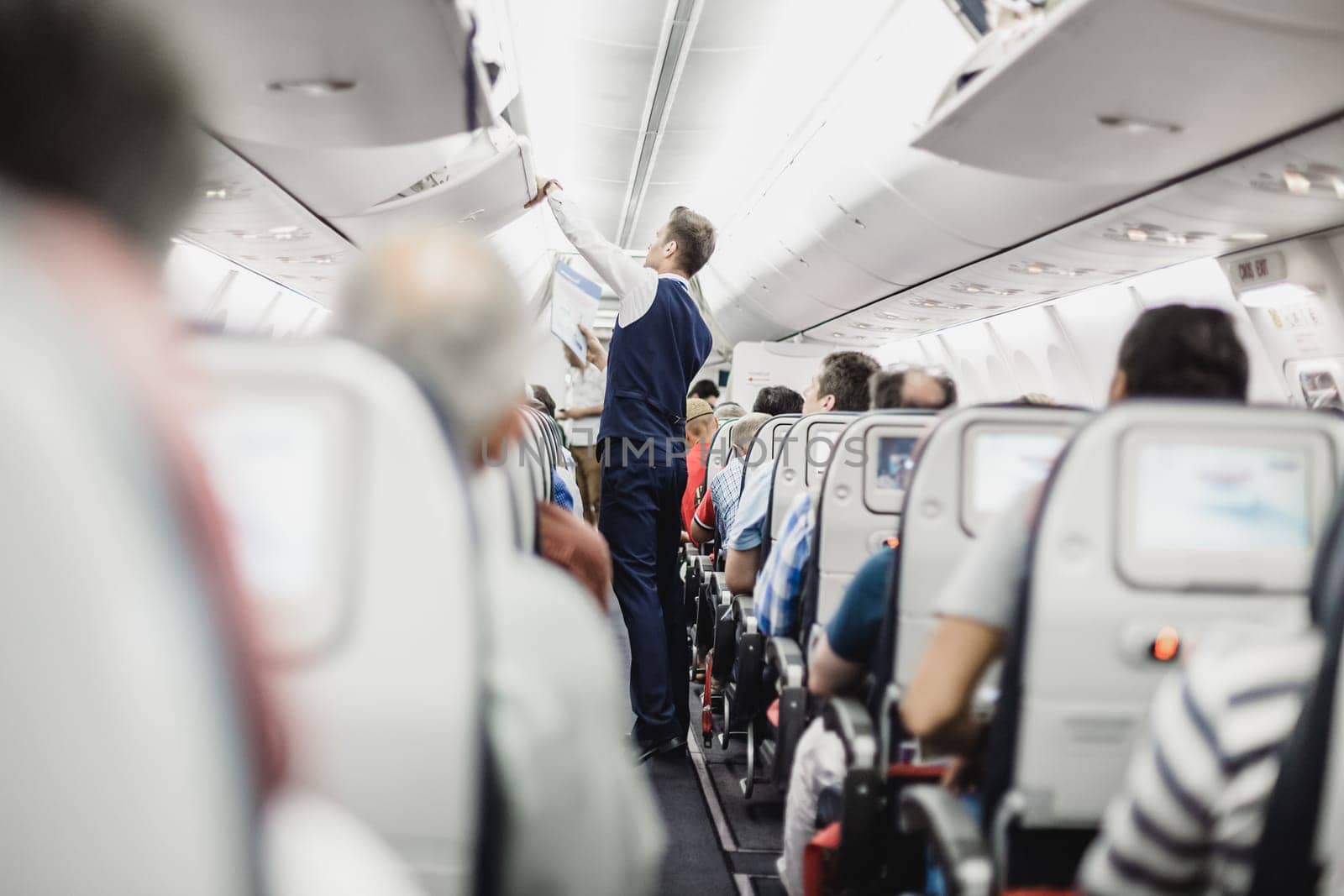 Interior of airplane with passengers on seats and steward in uniform closing overhead luggage compartment.