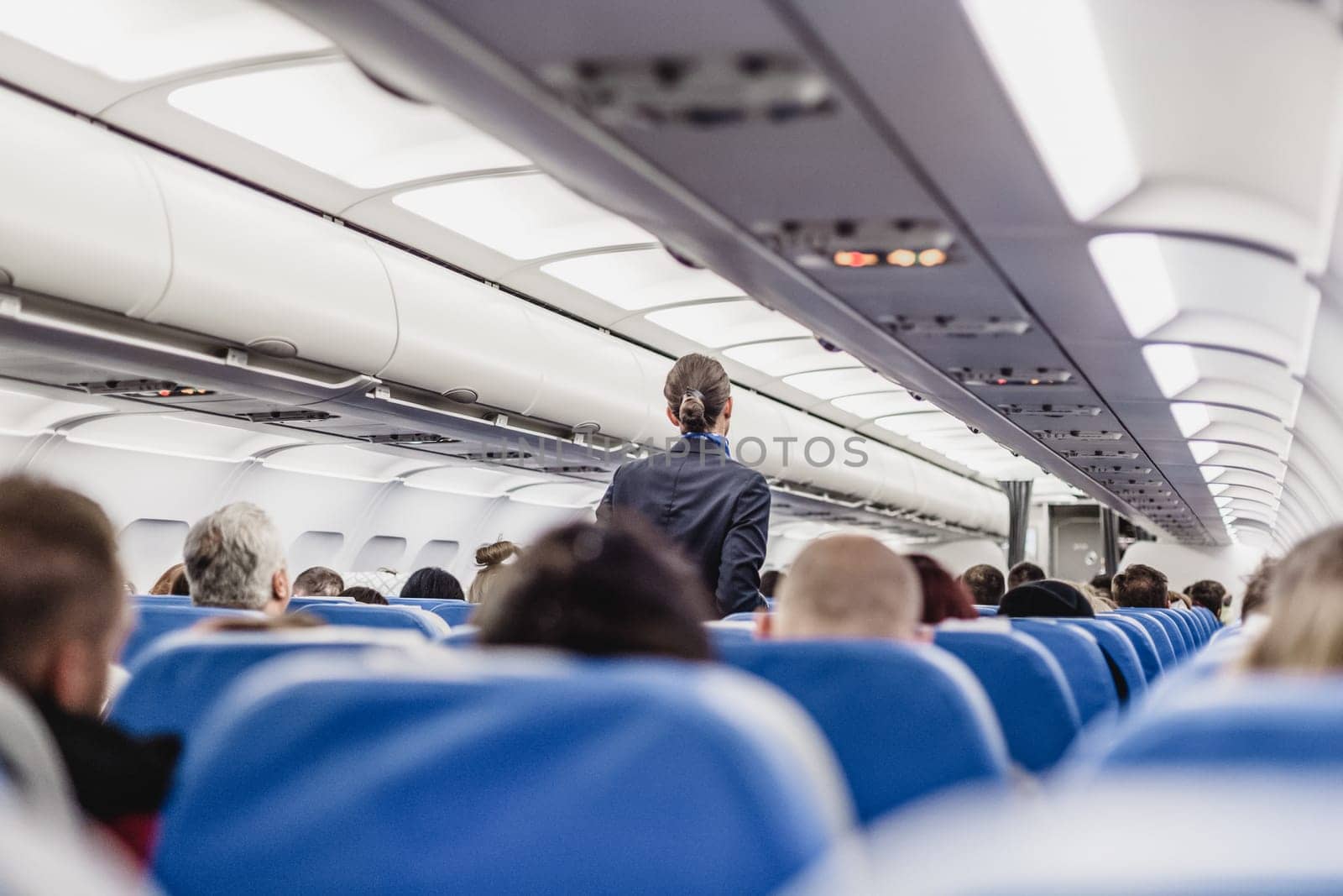 Interior of airplane with passengers on seats and stewardess walking the aisle.