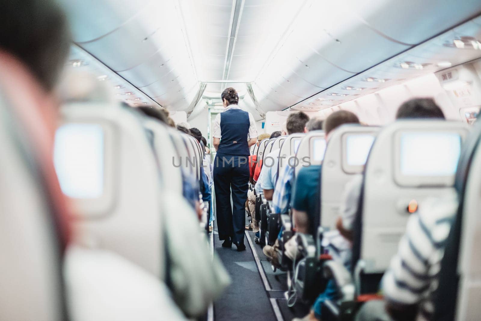 Interior of airplane with passengers on seats and stewardess in uniform walking the aisle.