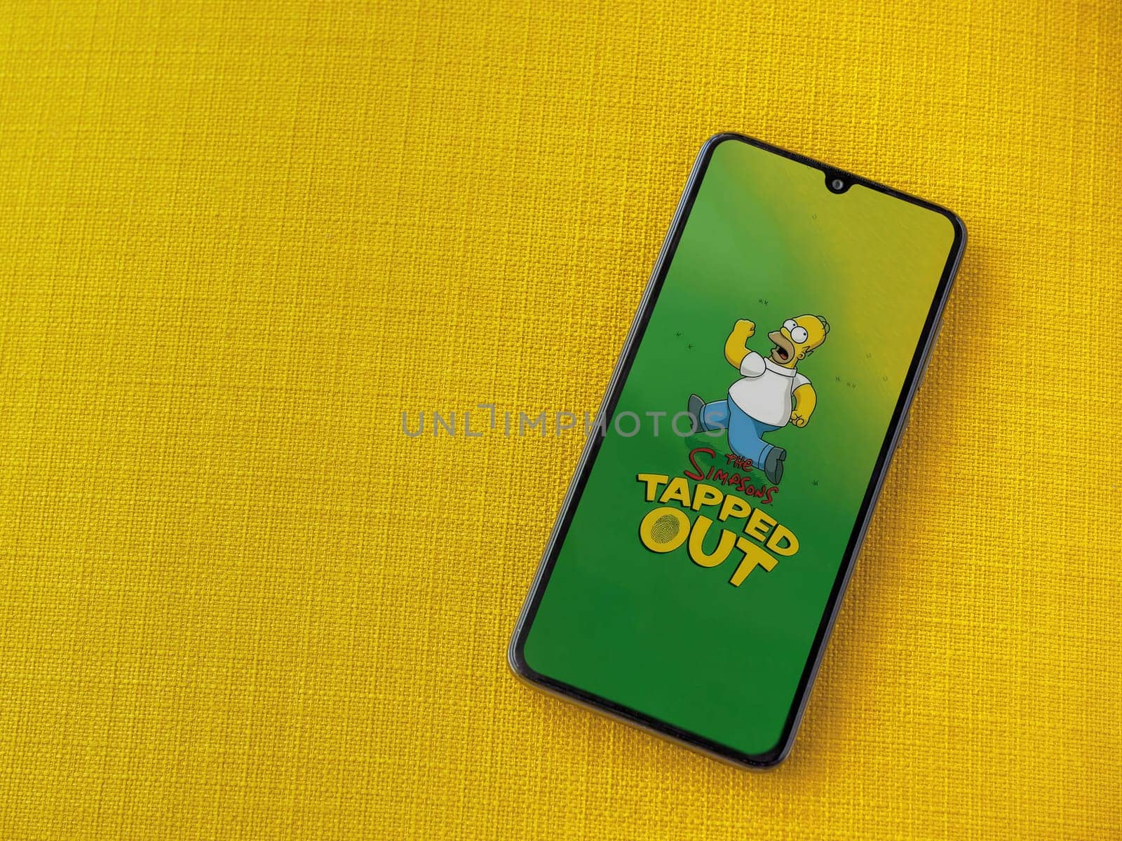Lod, Israel - July 16,2023: The Simpsons Tapped Out app launch screen on smartphone on yellow fabric background. Top view flat lay with copy space.