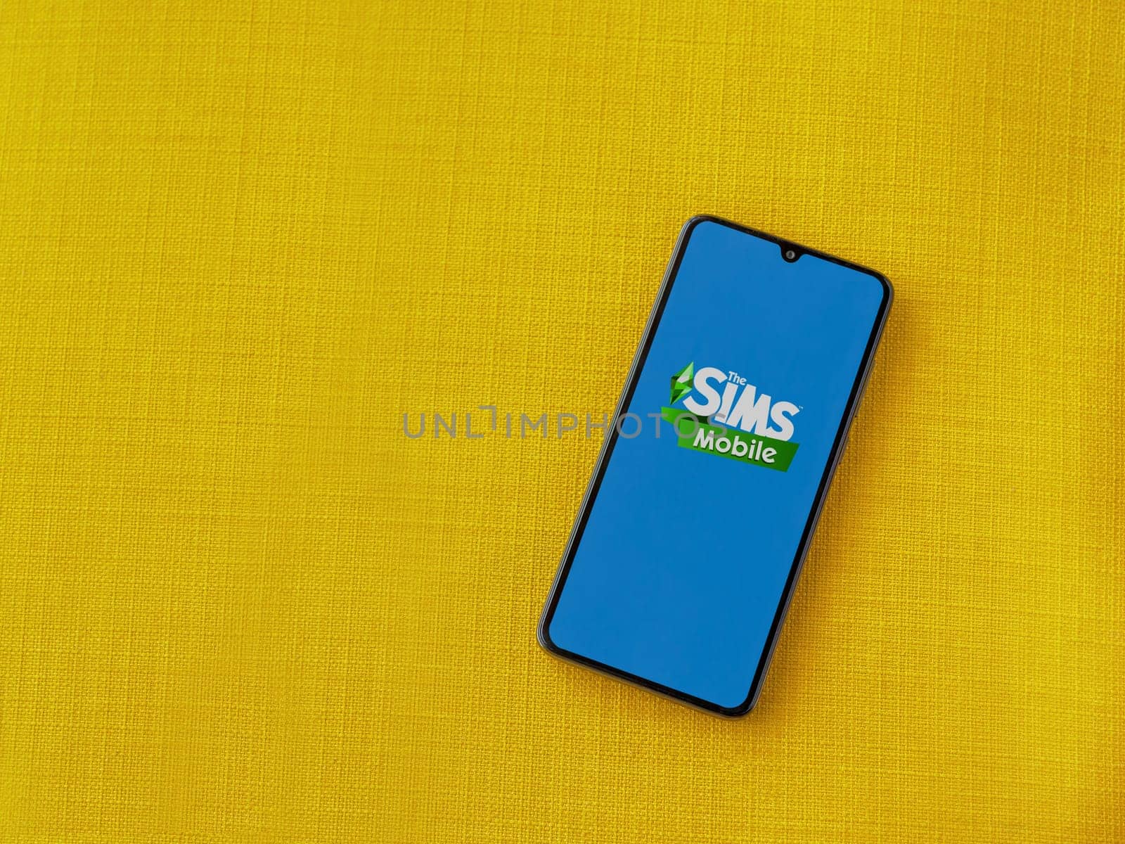 Lod, Israel - July 16,2023: The Sims Mobile app launch screen on smartphone on yellow fabric background. Top view flat lay with copy space.