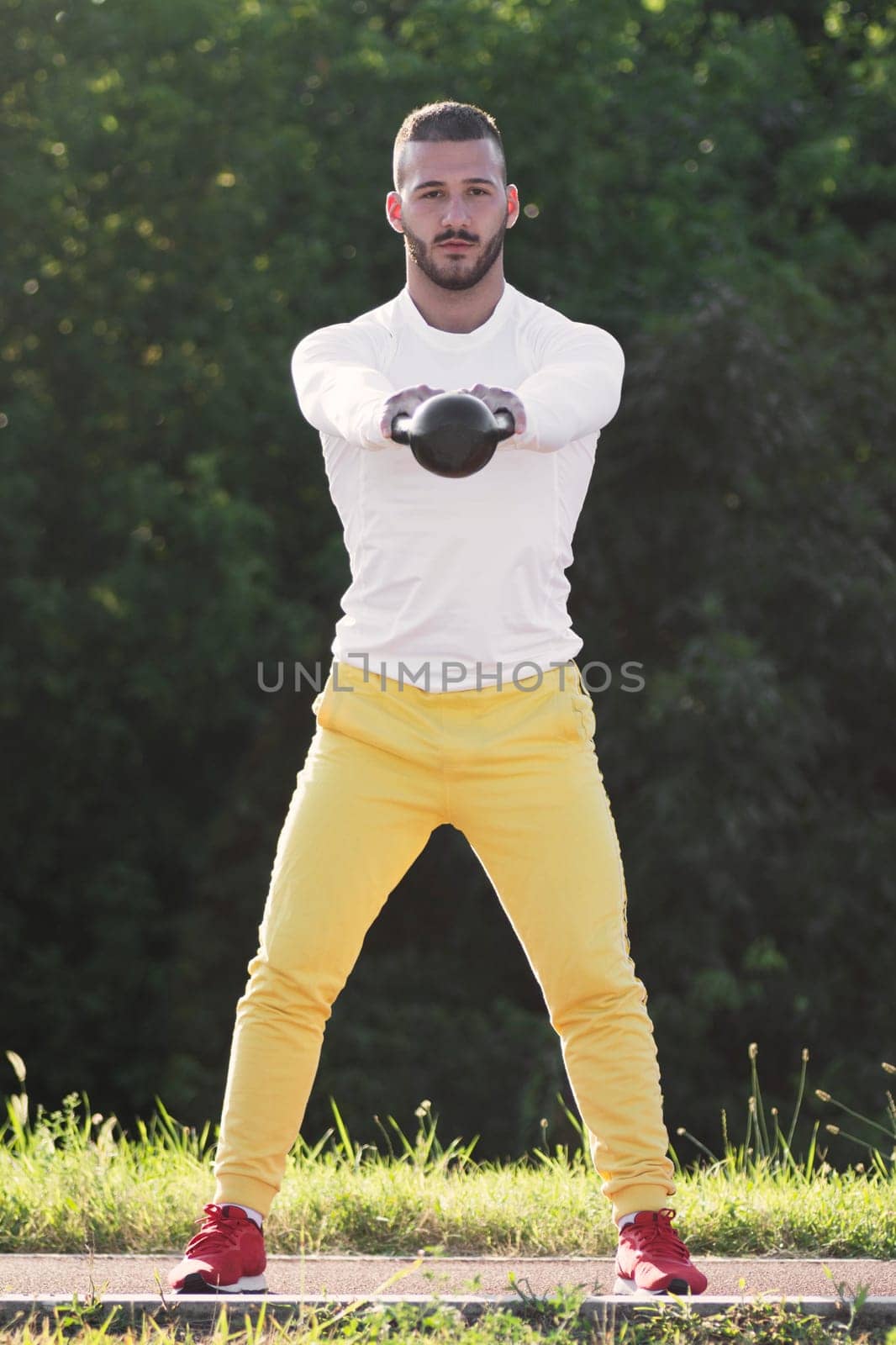 Front view of the sporty young guy is exercising with kettlebell sport equipment outdoors.