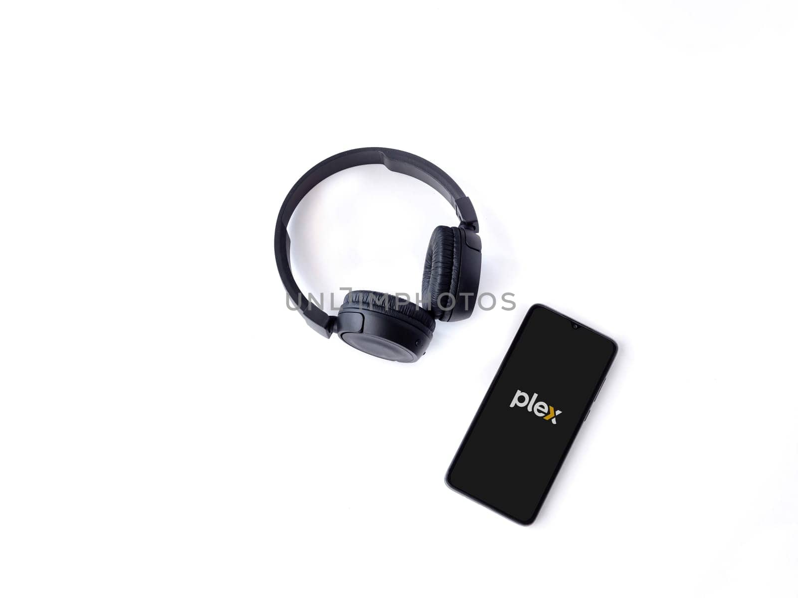 Lod, Israel - July 16,2023: Smartphone with Plex app launch screen and wireless headphones on white background. Top view flat lay with copy space.
