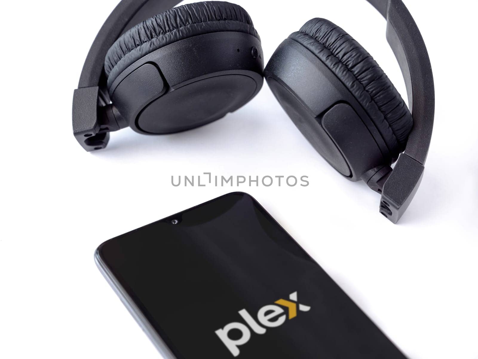 Lod, Israel - July 16,2023: Smartphone with Plex app launch screen and wireless headphones on white background. Top view flat lay with copy space.