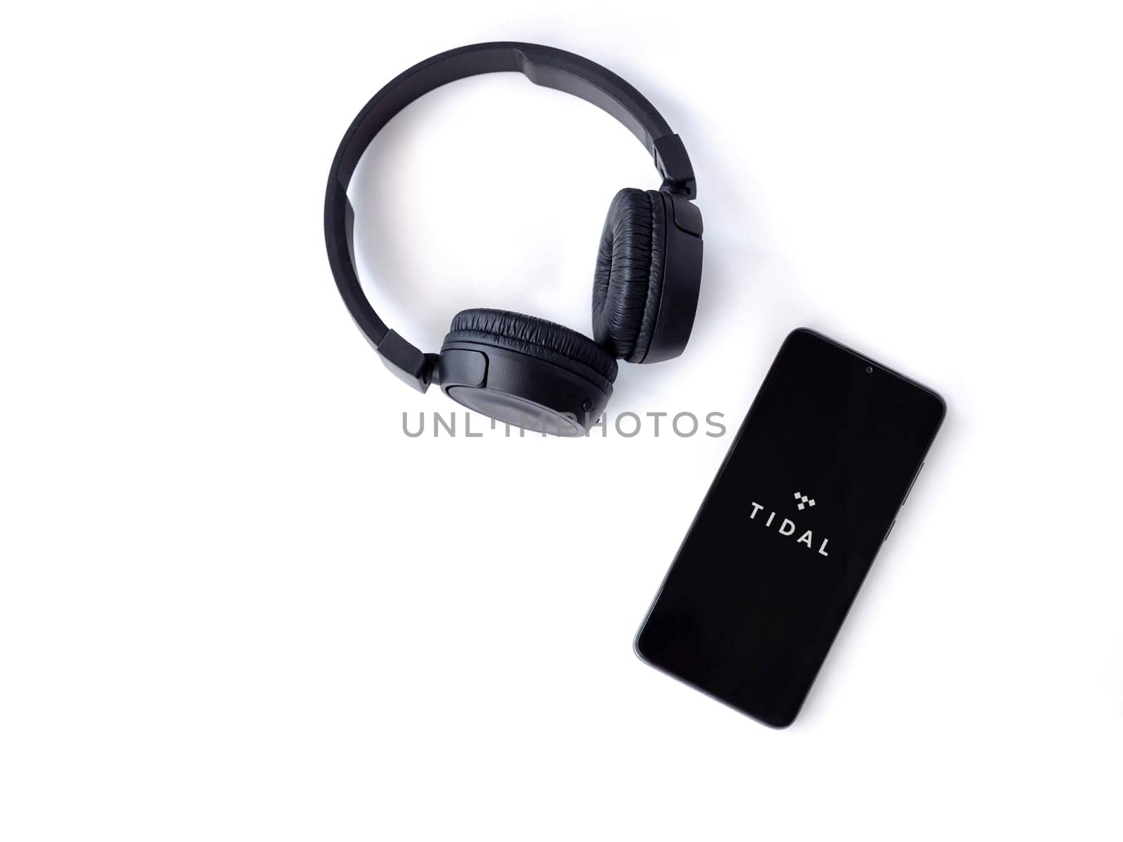 Lod, Israel - July 16,2023: Smartphone with TIDAL Music app launch screen and wireless headphones on white background. Top view flat lay with copy space.