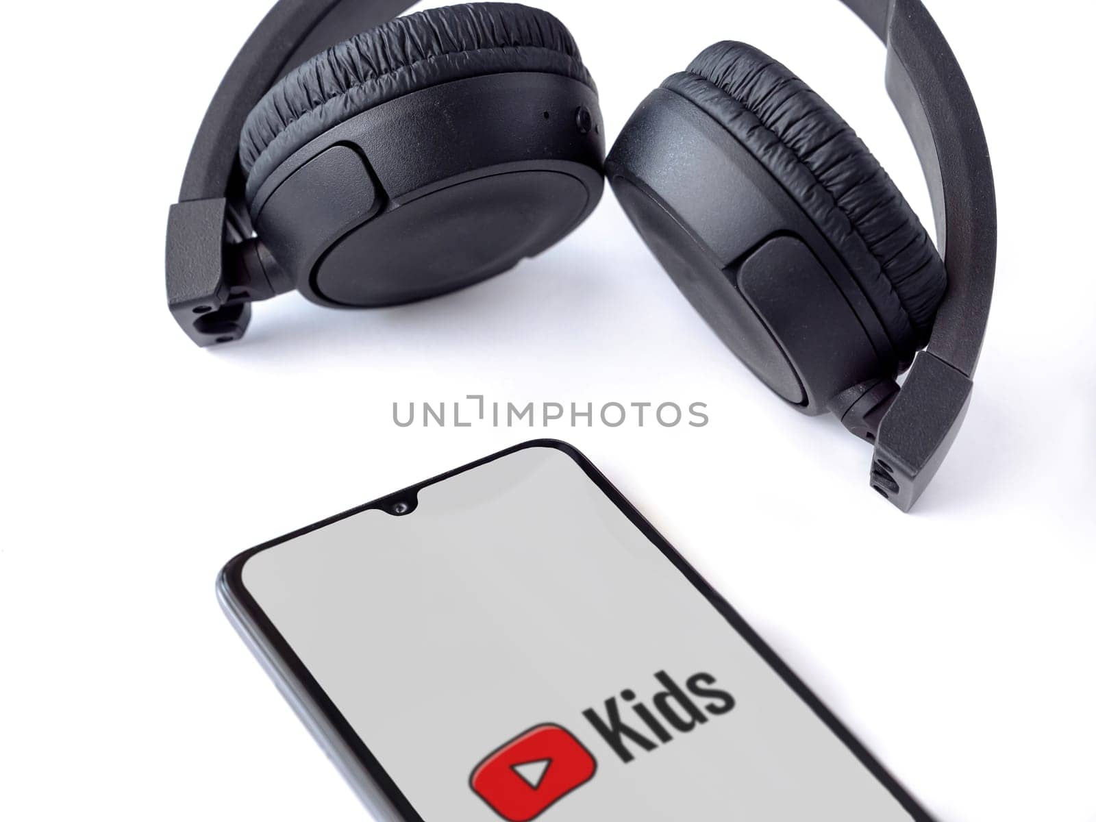 Lod, Israel - July 16,2023: Smartphone with YouTube Kids app launch screen and wireless headphones on white background. Top view flat lay with copy space.