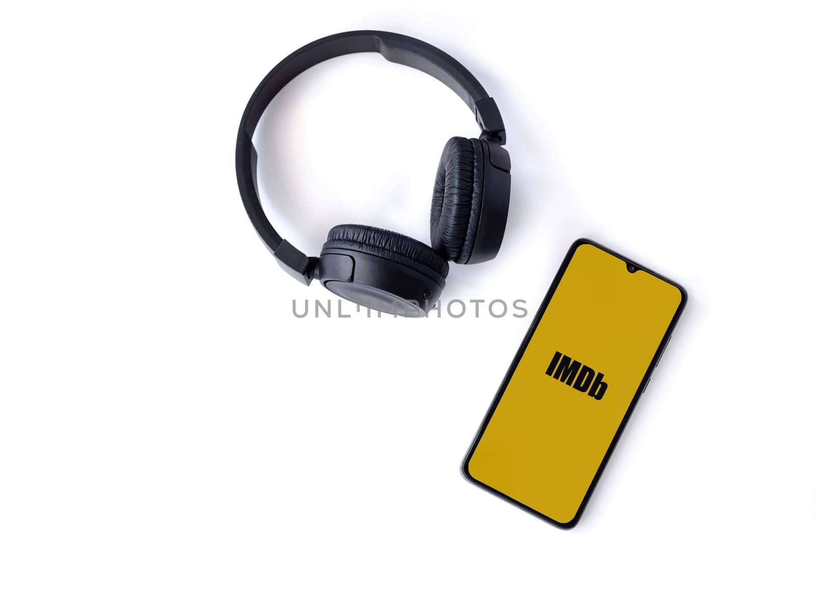 Lod, Israel - July 16,2023: Smartphone with IDMb app launch screen and wireless headphones on white background. Top view flat lay with copy space.