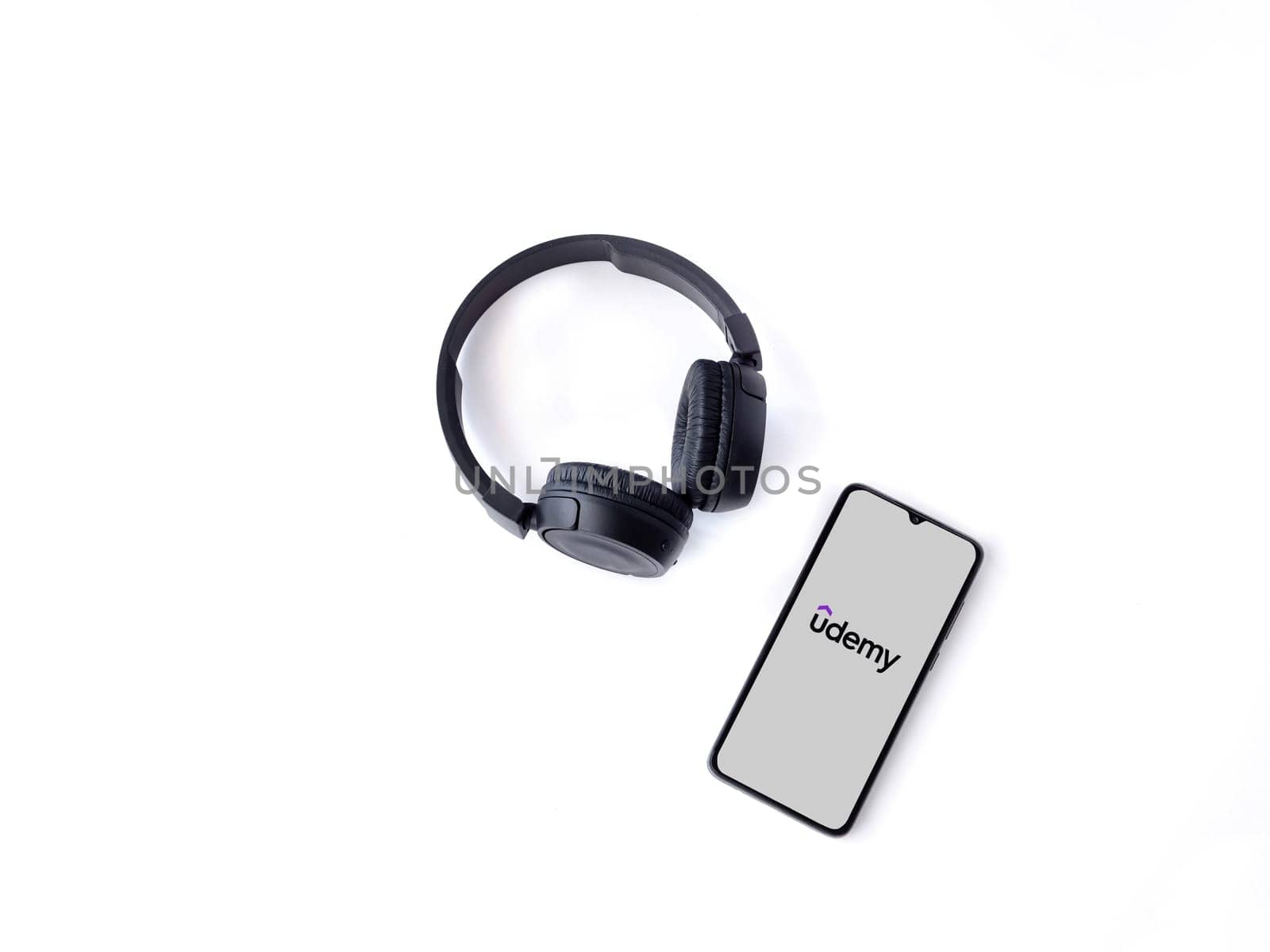 Lod, Israel - July 16,2023: Smartphone with Udemy app launch screen and wireless headphones on white background. Top view flat lay with copy space.