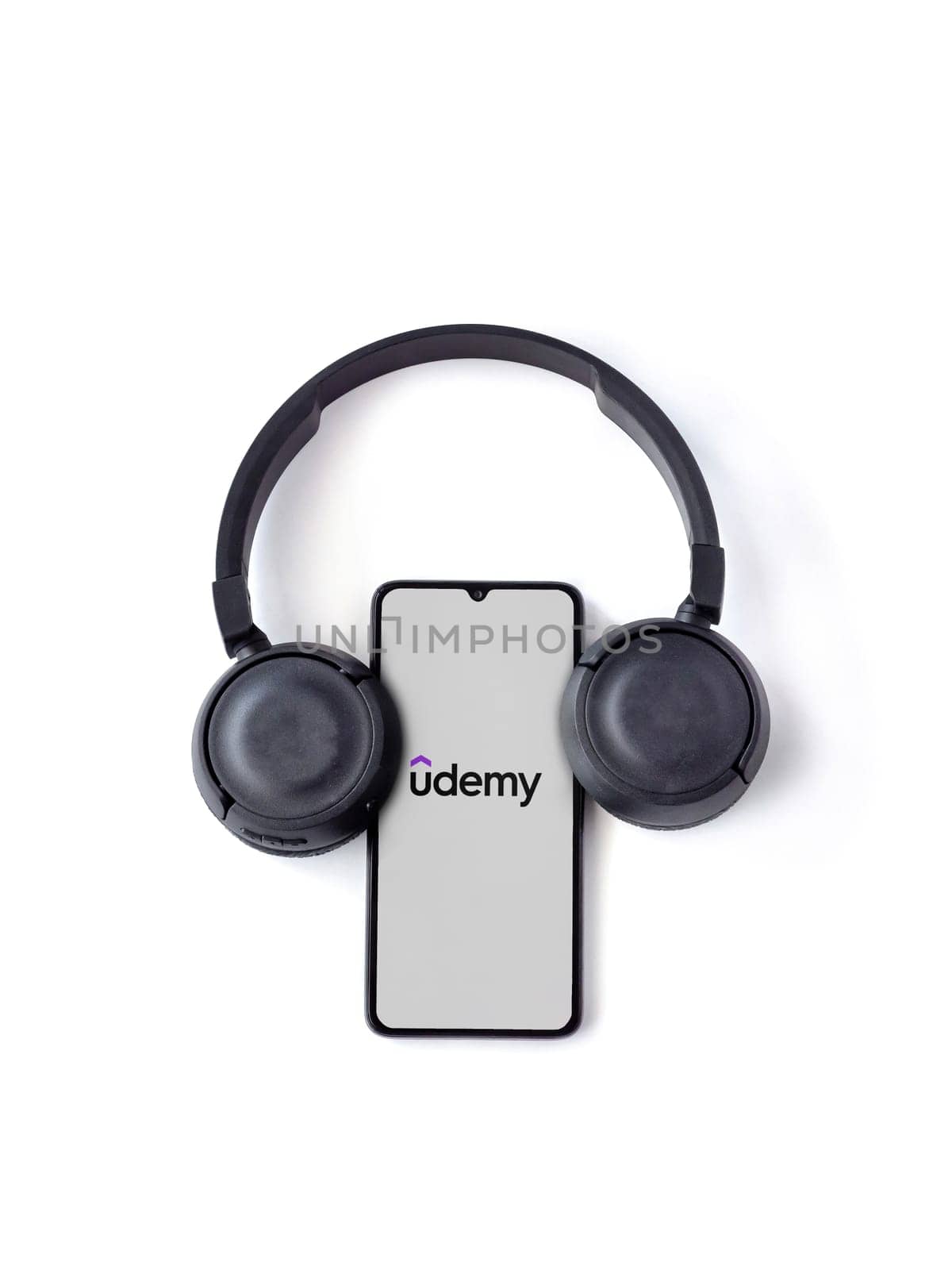 Lod, Israel - July 16,2023: Smartphone with Udemy app launch screen and wireless headphones on white background. Top view flat lay with copy space.