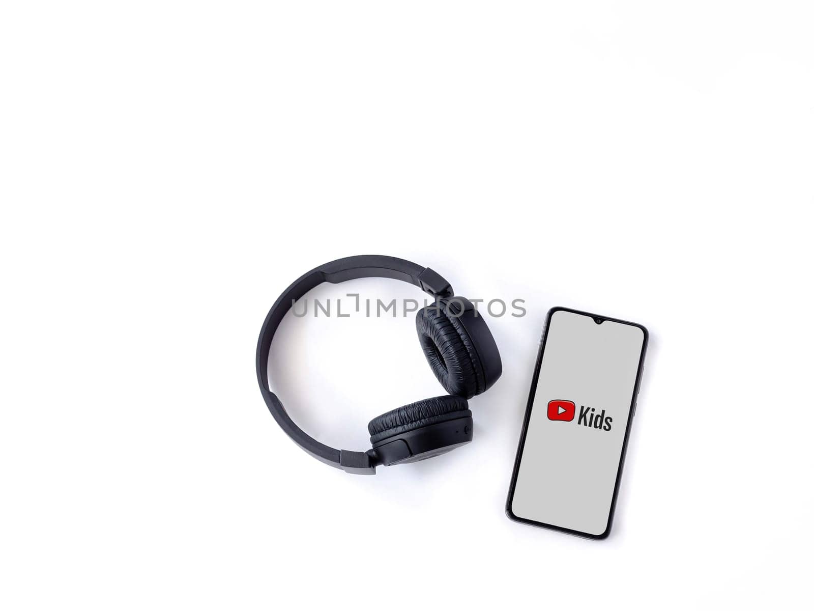 Lod, Israel - July 16,2023: Smartphone with YouTube Kids app launch screen and wireless headphones on white background. Top view flat lay with copy space.