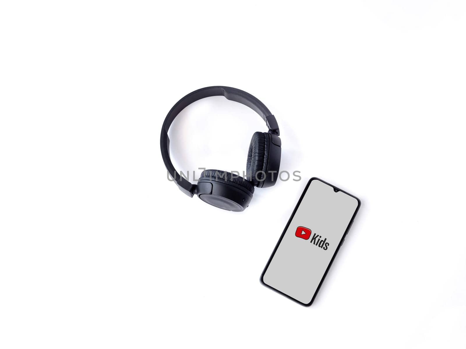 Lod, Israel - July 16,2023: Smartphone with YouTube Kids app launch screen and wireless headphones on white background. Top view flat lay with copy space.