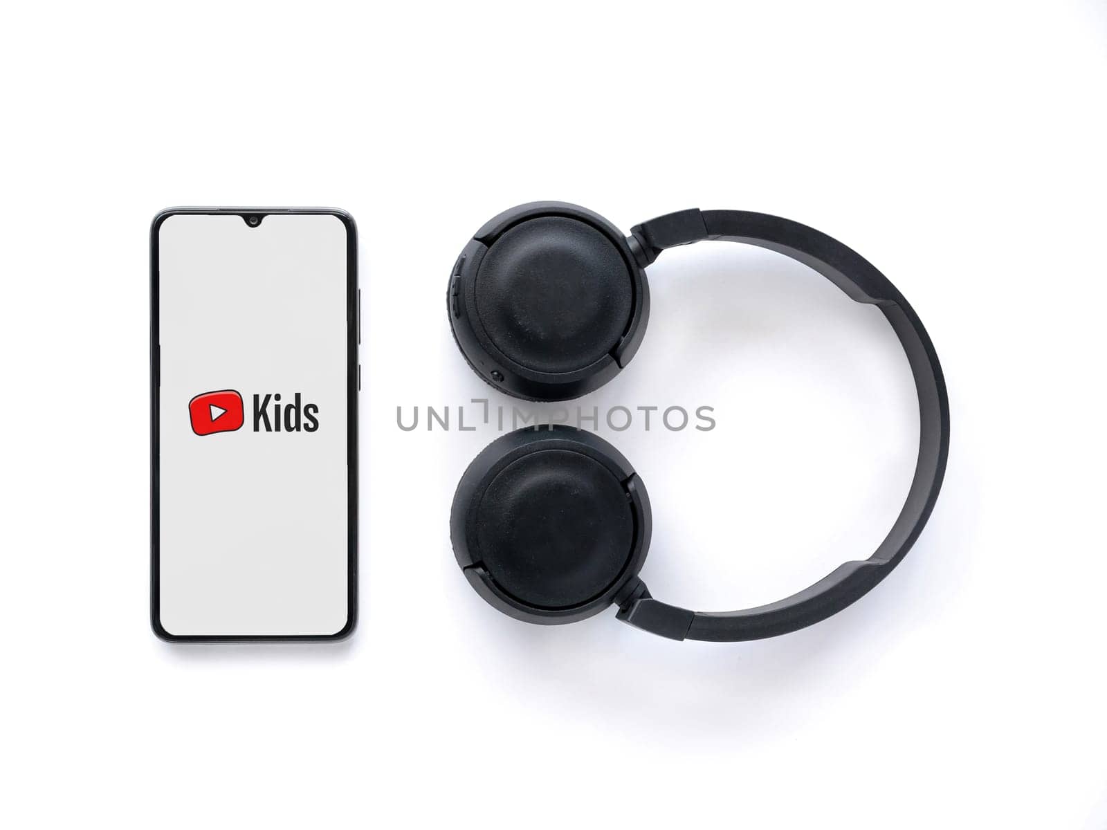 Lod, Israel - July 16,2023: Smartphone with YouTube Kids app launch screen and wireless headphones on white background. Top view flat lay with copy space.